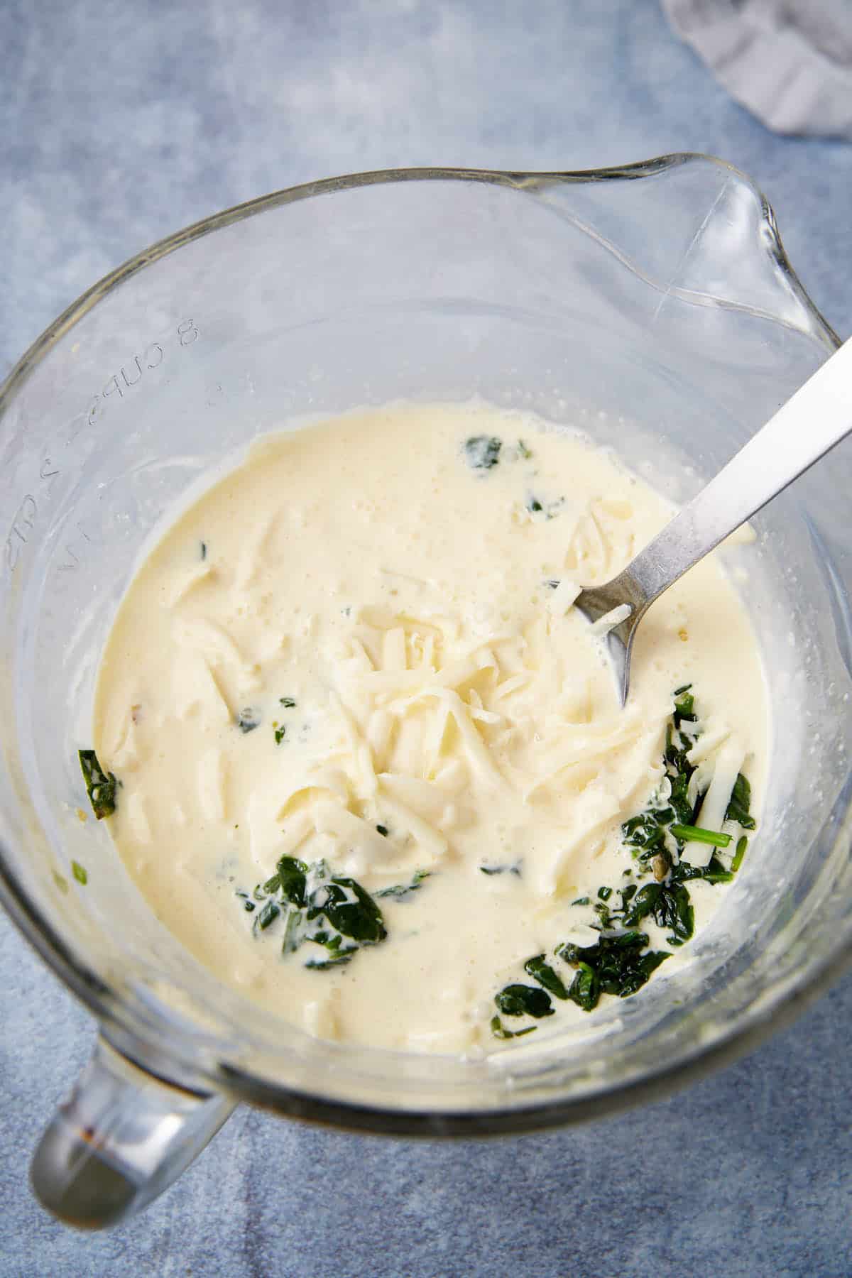 A glass mixing bowl filled with a creamy white mixture, shredded cheese, and chopped spinach—perfect for making Prosciutto Egg Cups—sits on a light blue surface with a metal spoon resting inside.