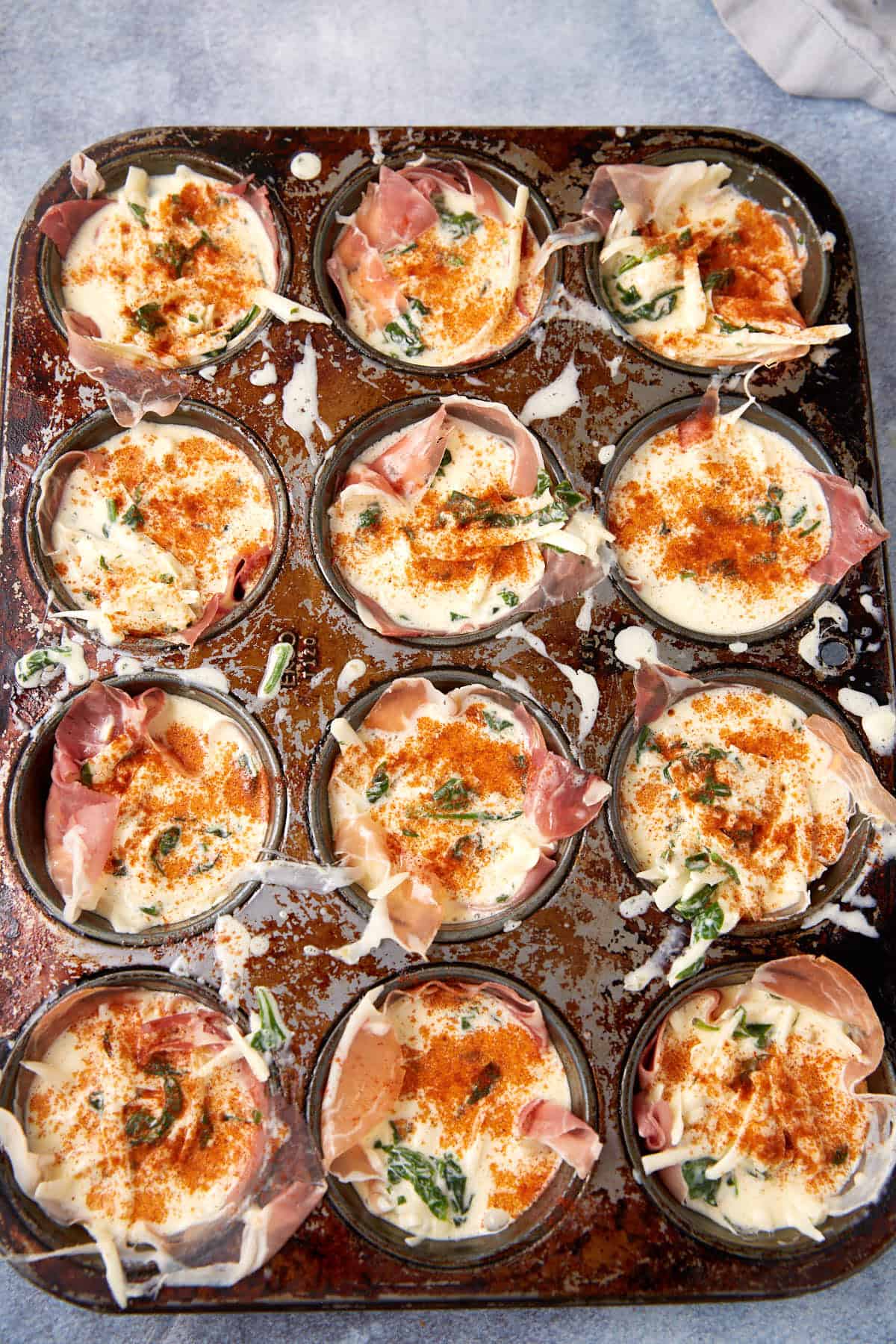 A muffin tin holds twelve uncooked Prosciutto Egg Cups, each lined with prosciutto, spinach, and cheese, then dusted with paprika on a rustic baking tray. Some filling has spilled onto the metal tray.