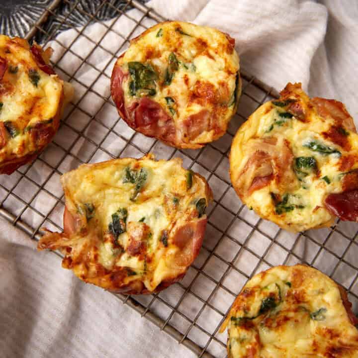 Five baked Prosciutto Egg Cups with spinach and ham sit on a cooling rack over a cream-colored cloth. The muffins are golden brown, showcasing visible bits of green and pink ingredients.