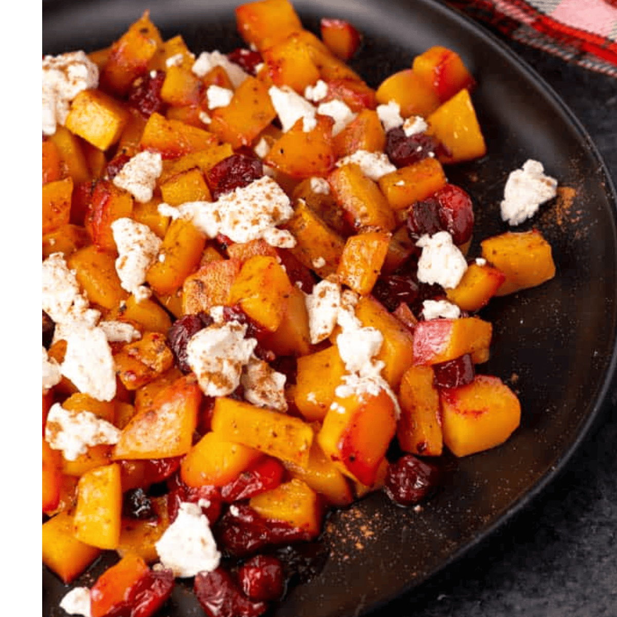 A black plate with roasted Butternut Squash with Cranberries and Feta, garnished with spices. Dried cranberries and crumbled cheese add color, while a red and white cloth is partially visible in the corner.