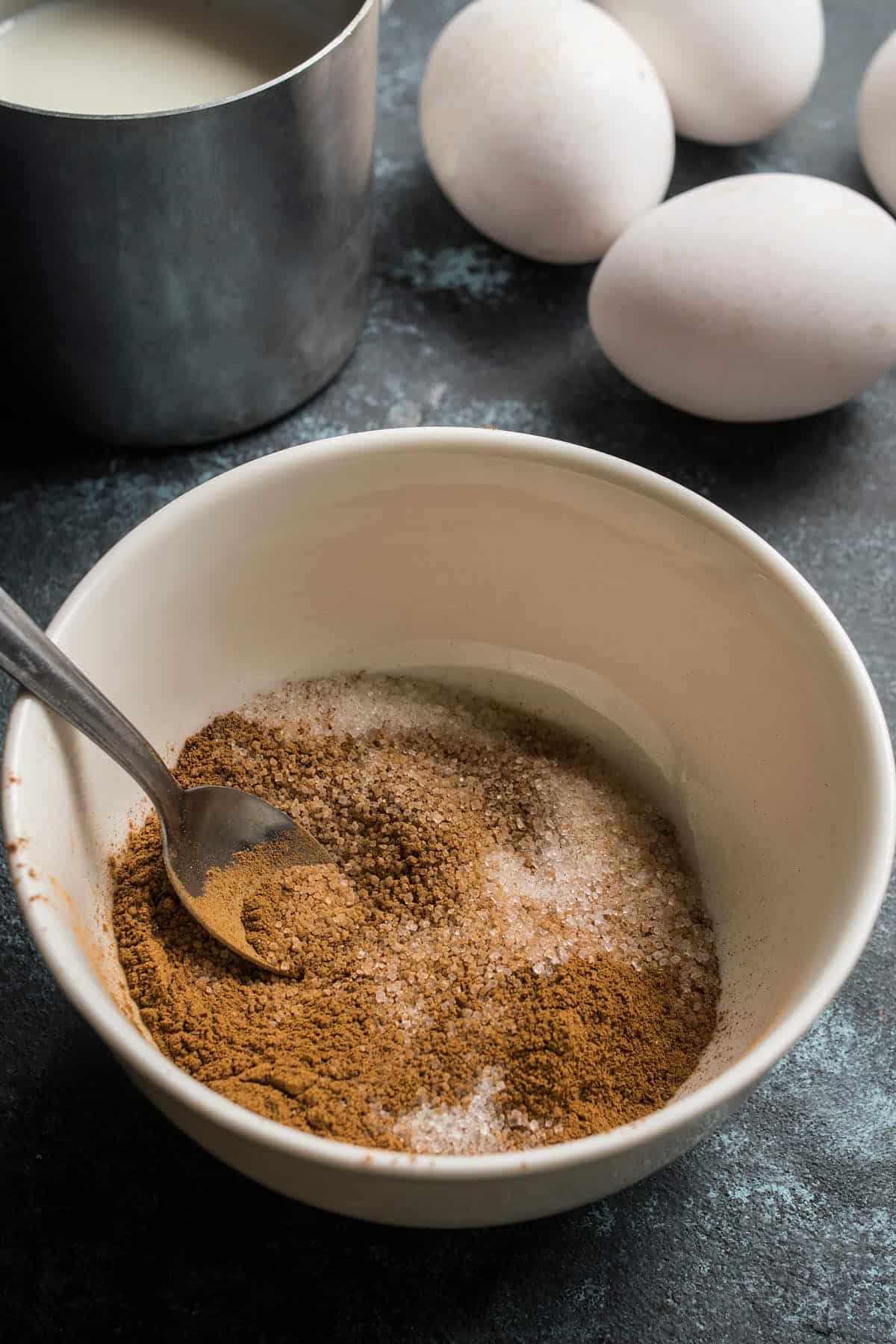 A bowl with a spoon mixing cinnamon and sugar for a German apple pancake, next to three white eggs and a metal cup containing milk on a dark surface.
