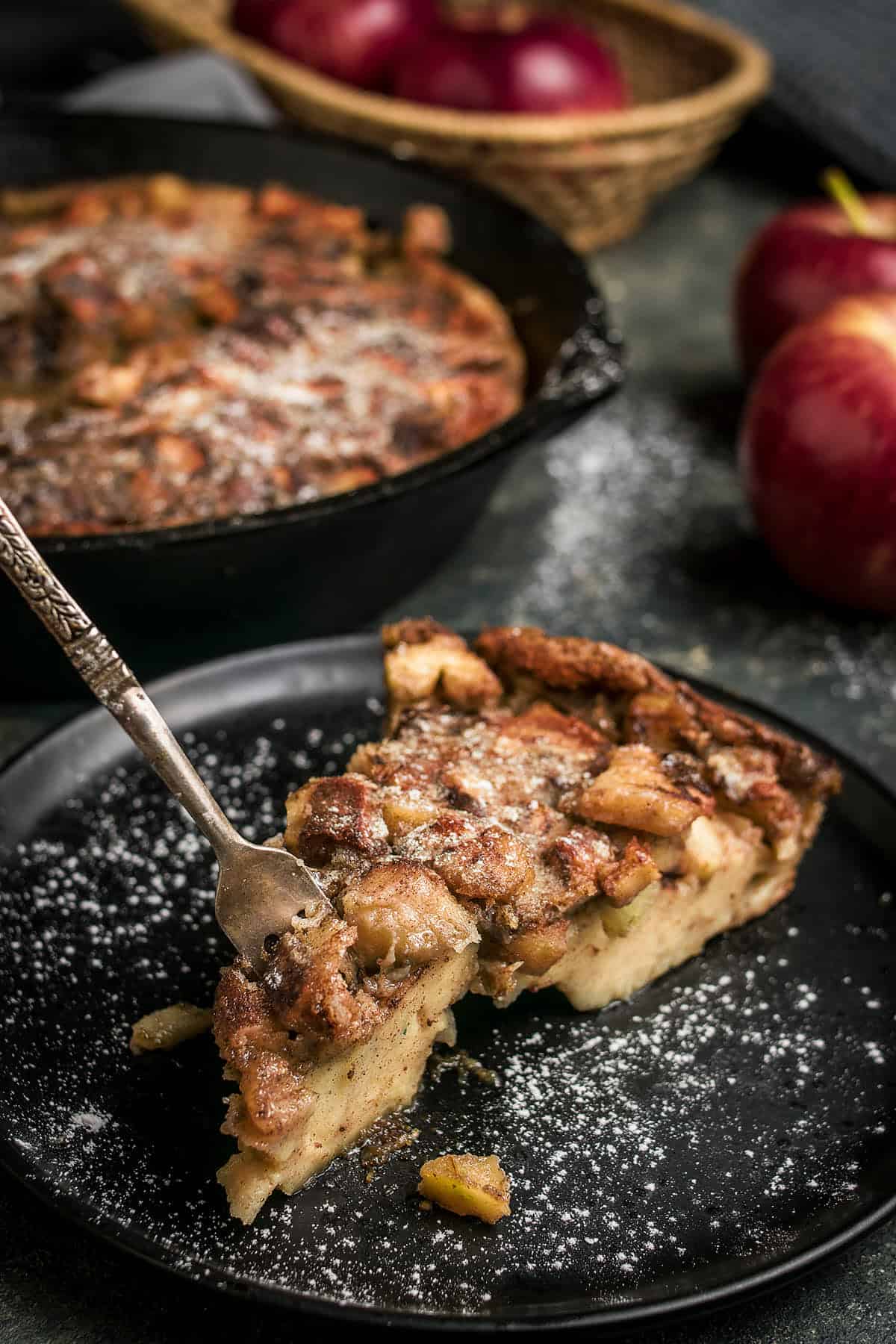 A slice of apple cinnamon bread pudding, reminiscent of a German apple pancake, is being lifted with a fork from a black plate dusted with powdered sugar. In the background, a cast iron skillet holds more pudding alongside fresh red apples.