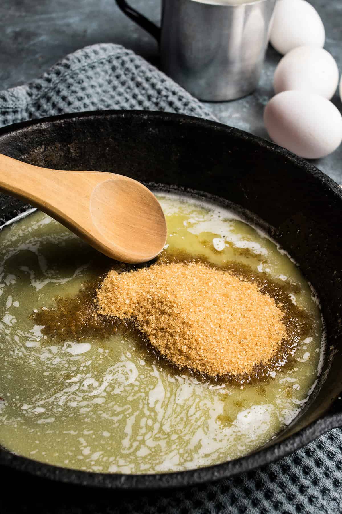 A wooden spoon rests in a black cast iron skillet with melted butter and a mound of brown sugar—perfect beginnings for a German apple pancake. Three white eggs and a metal cup sit in the background on a textured blue cloth.