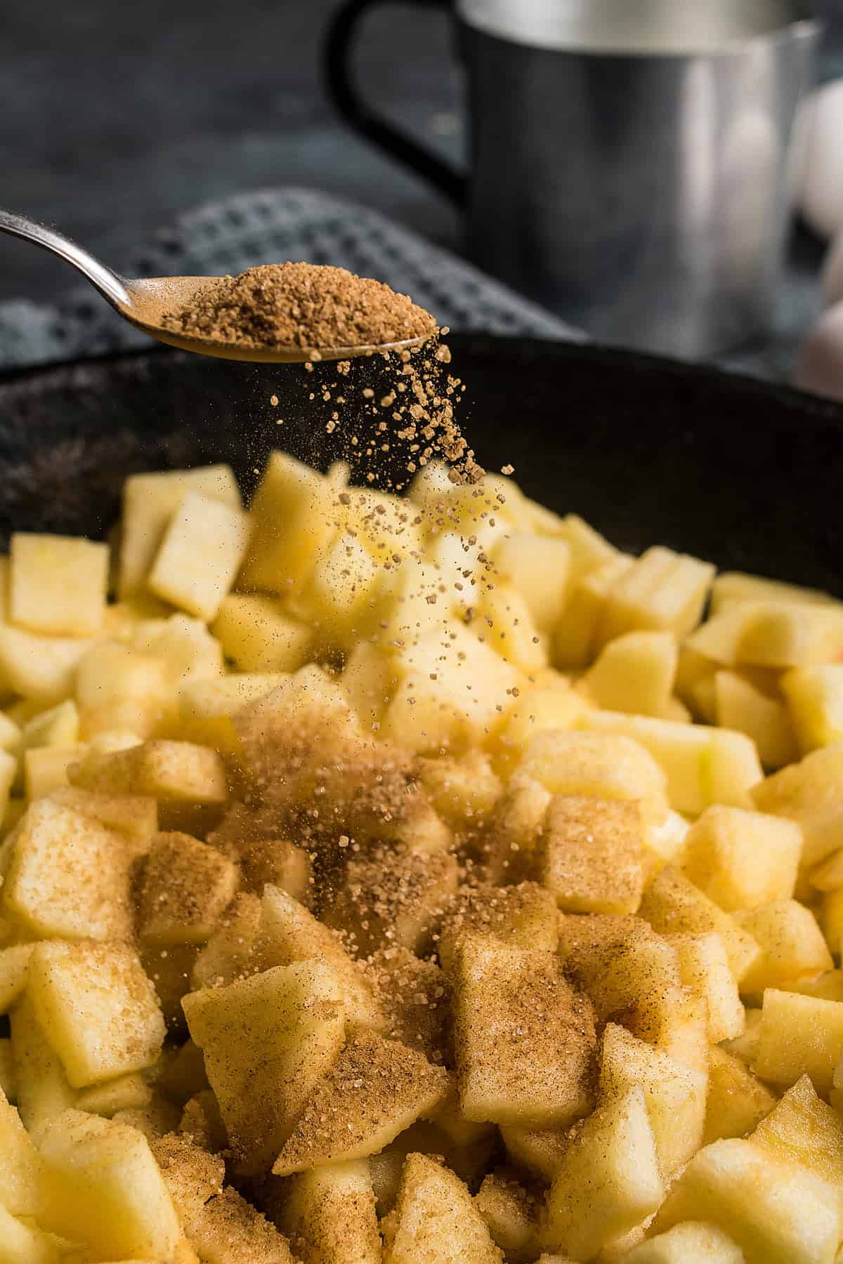 A spoon sprinkles brown sugar over a pile of diced apples in a skillet, hinting at the first steps of making a delicious German apple pancake; a metal cup and towel are blurred in the background.