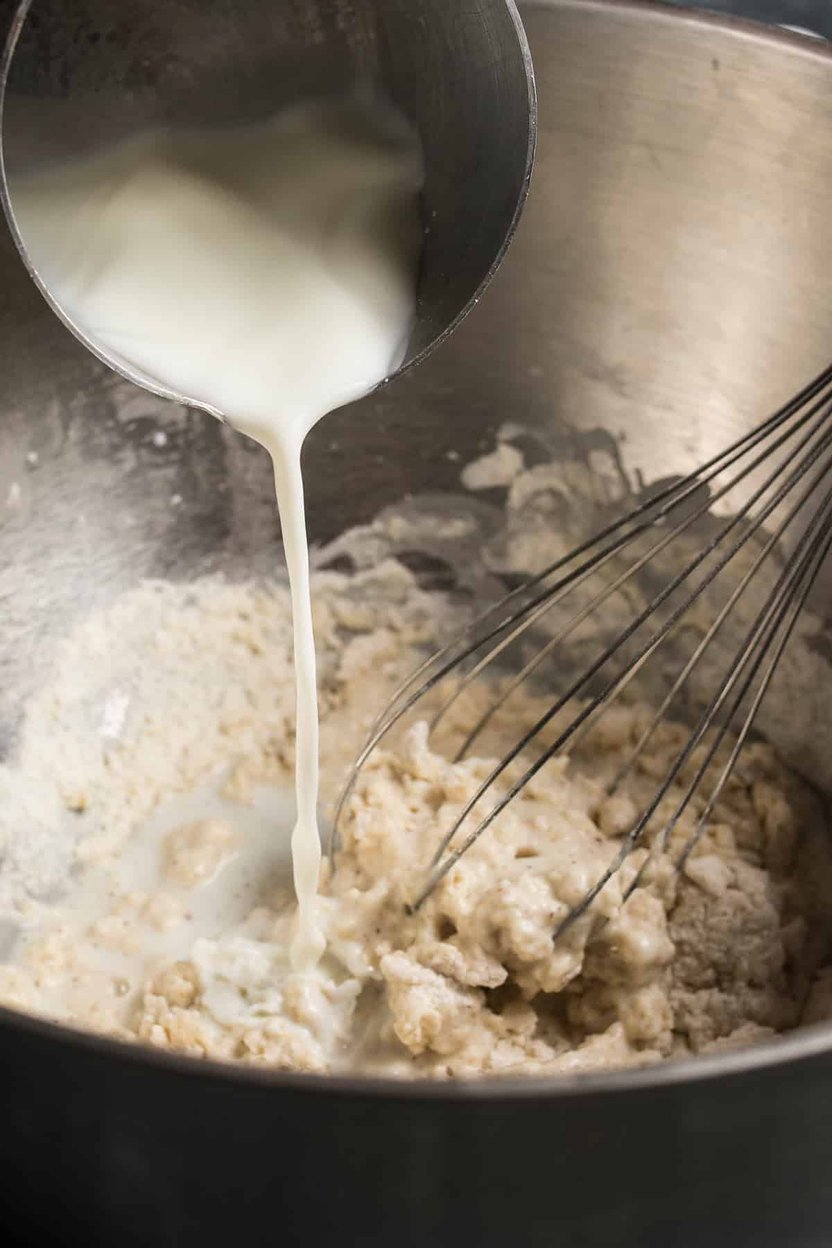 A close-up of milk being poured from a metal cup into a mixing bowl with flour and dough, next to a metal whisk—just the start of creating the perfect batter for a classic German apple pancake.