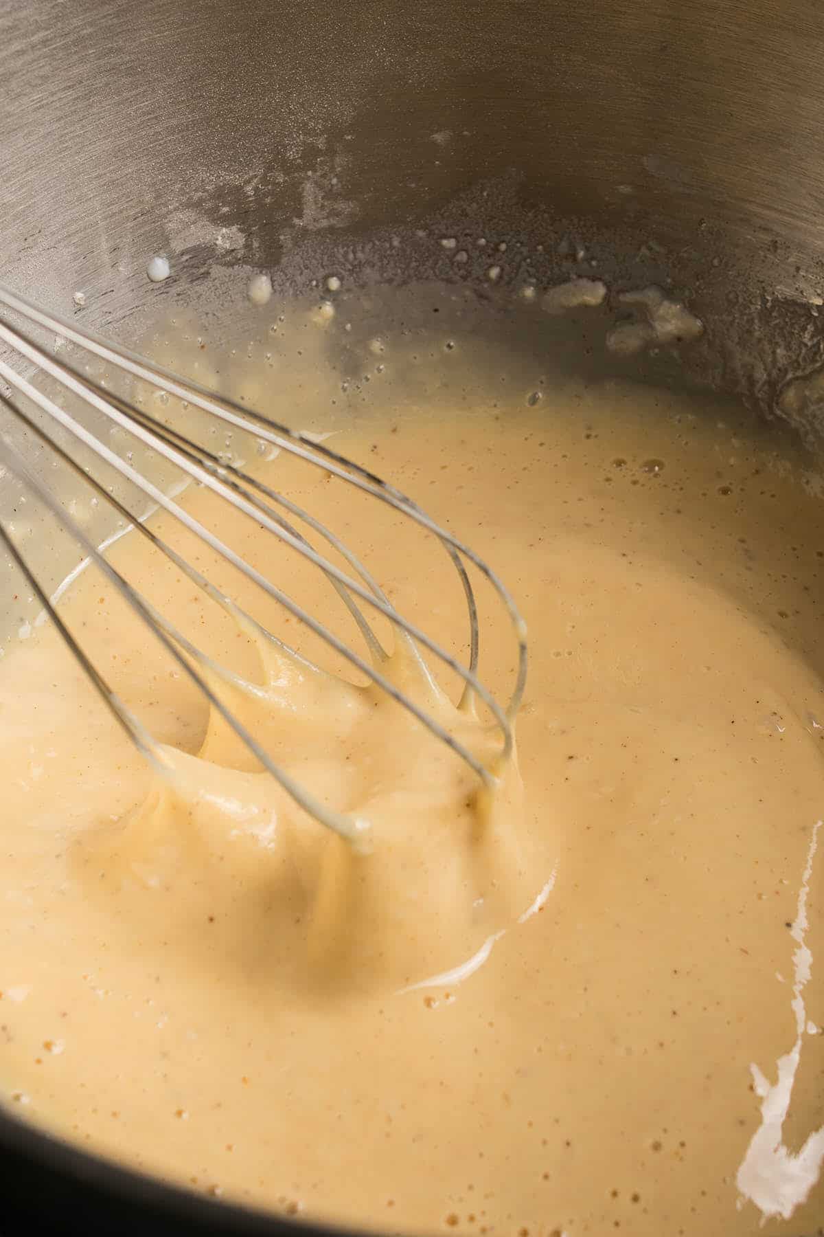A metal whisk mixing a thick, creamy German apple pancake batter in a stainless steel bowl. Some batter clings to the whisk, and there are small droplets and bubbles on the sides of the bowl.