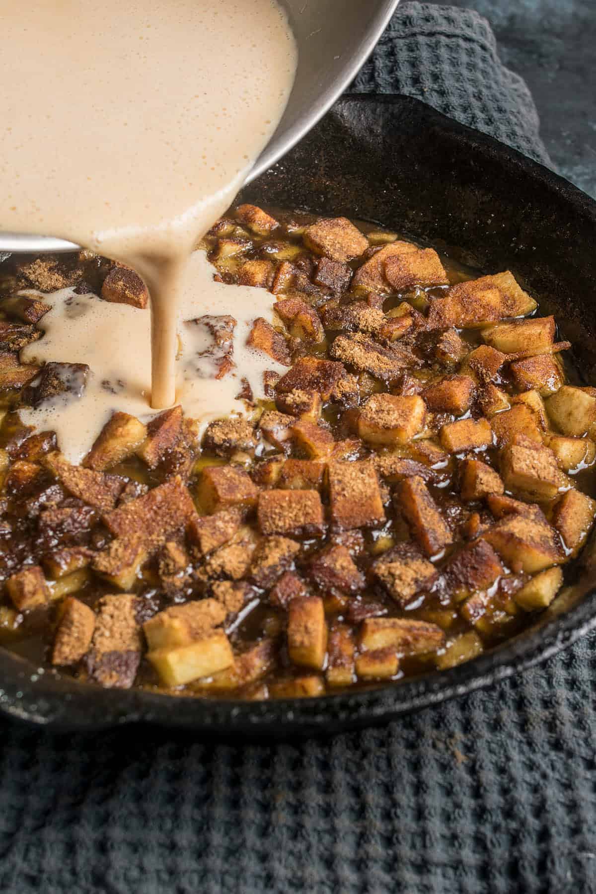 A creamy mixture is being poured from a metal bowl onto diced apples and bread cubes in a cast iron skillet, creating the base for a delicious German apple pancake, all resting on a textured dark cloth.