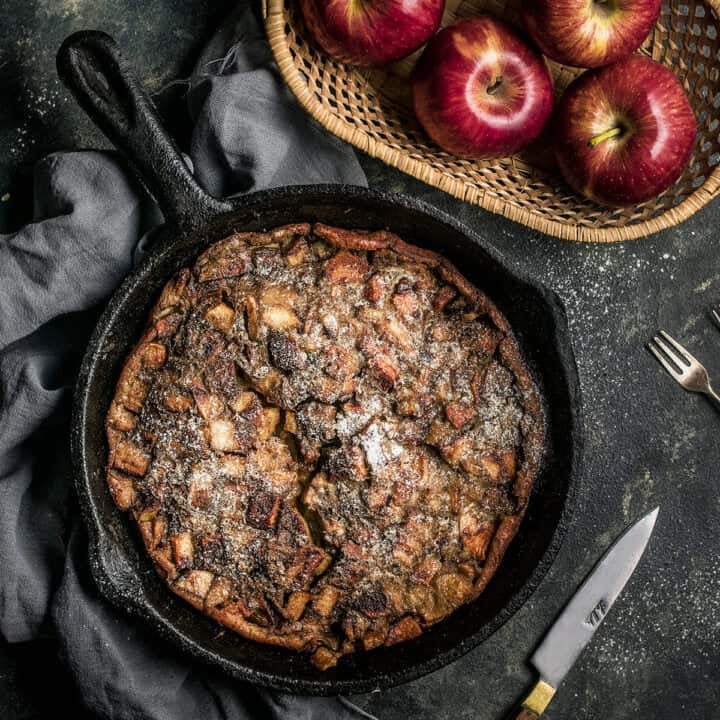 A skillet filled with a baked German apple pancake dusted with powdered sugar sits on a dark cloth beside a basket of red apples, with a fork and knife nearby on the dark tabletop.
