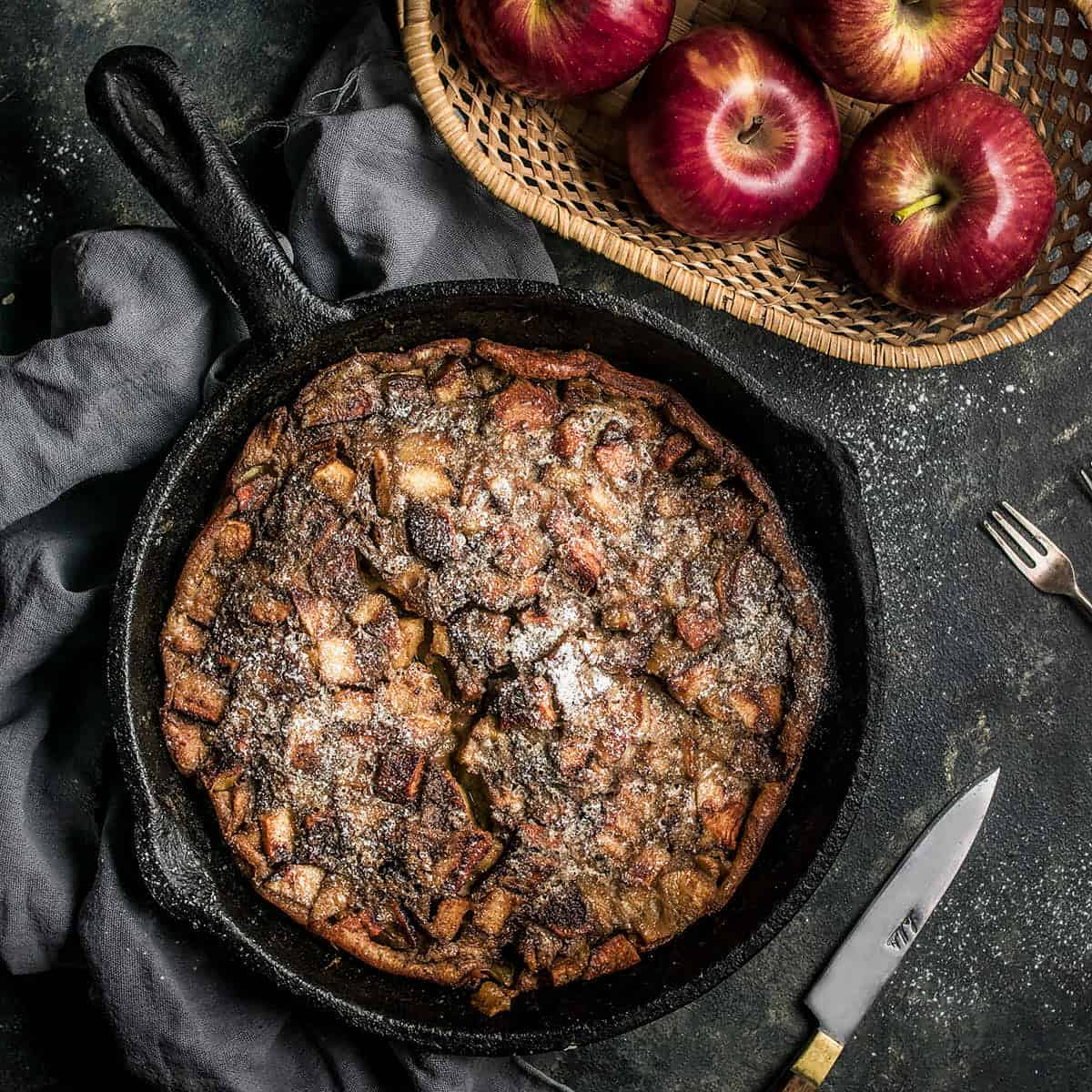 A skillet filled with a baked German apple pancake dusted with powdered sugar sits on a dark cloth beside a basket of red apples, with a fork and knife nearby on the dark tabletop.