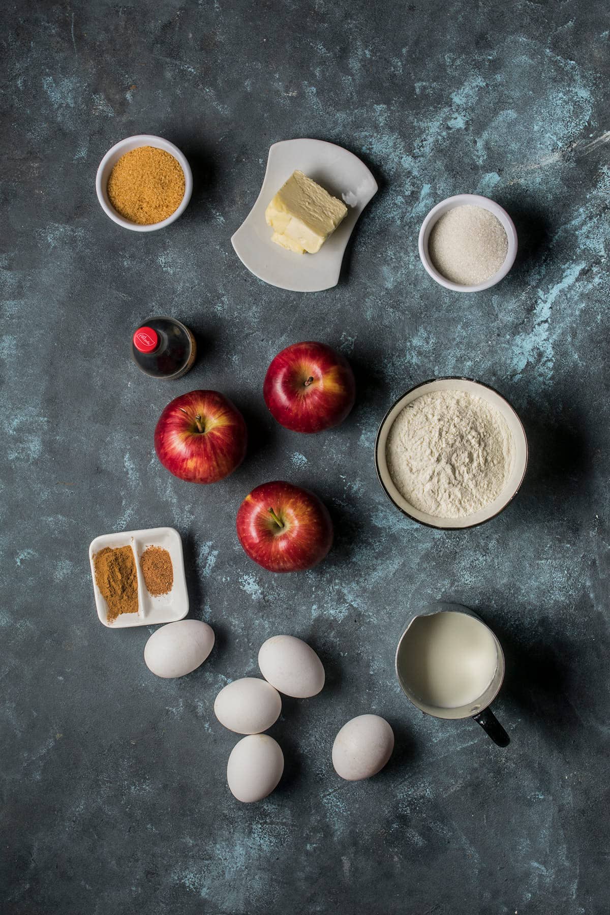 Flat lay of baking ingredients for a German apple pancake on a dark surface: three red apples, four eggs, a cup of flour, butter, milk, bowls of white and brown sugar, vanilla extract, cinnamon, and nutmeg.