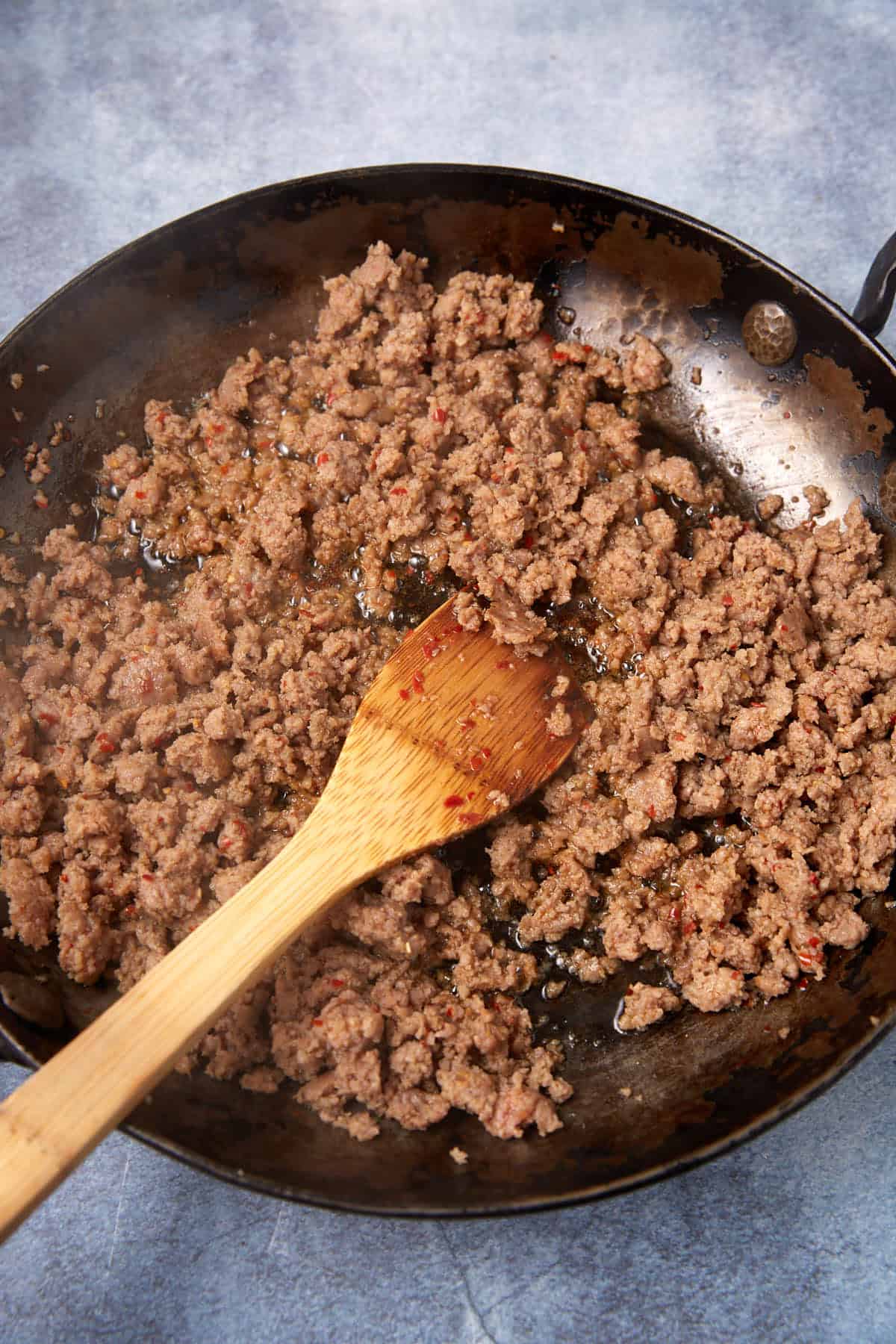 Ground meat is cooking in a black skillet, being stirred with a wooden spoon. The browned, crumbly texture contrasts with bits of seasoning—an inviting sight on the gray surface, much like a German apple pancake fresh from the pan.