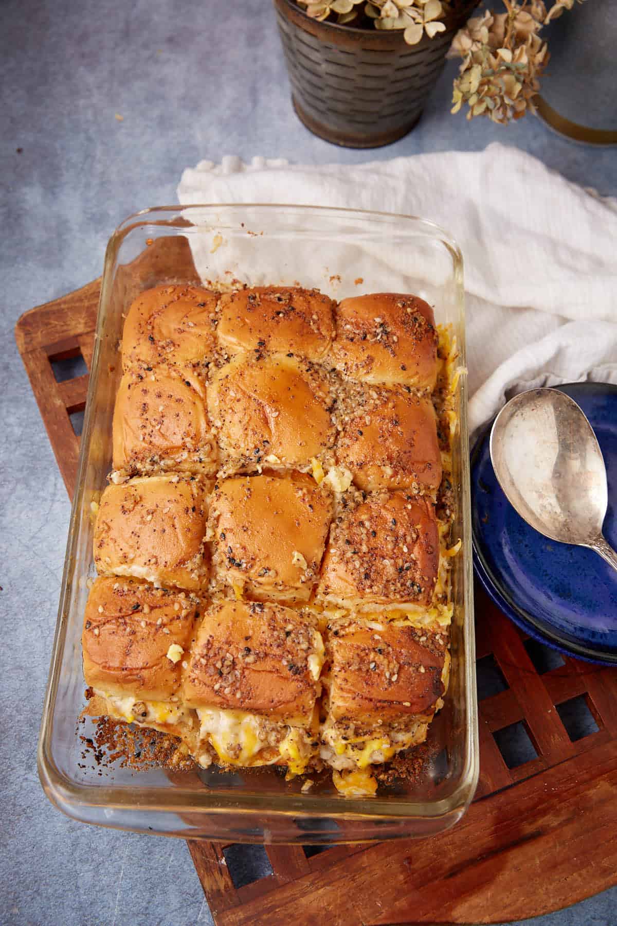 A glass baking dish filled with golden baked slider sandwiches, reminiscent of a German apple pancake’s warmth, sits on a wooden tray next to stacked blue plates, a spoon, and a white cloth napkin. Dried flowers are in the background.