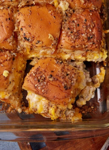 A glass baking dish filled with breakfast slider sandwiches, featuring soft buns topped with seasoning and stuffed with scrambled eggs, cheese, and sausage—reminiscent of a savory twist on a German apple pancake—with one corner slider partially removed.