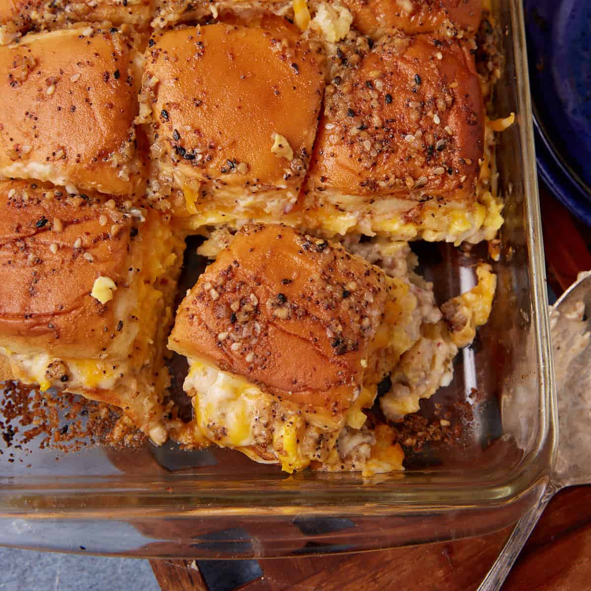 A glass baking dish filled with breakfast slider sandwiches, featuring soft buns topped with seasoning and stuffed with scrambled eggs, cheese, and sausage—reminiscent of a savory twist on a German apple pancake—with one corner slider partially removed.