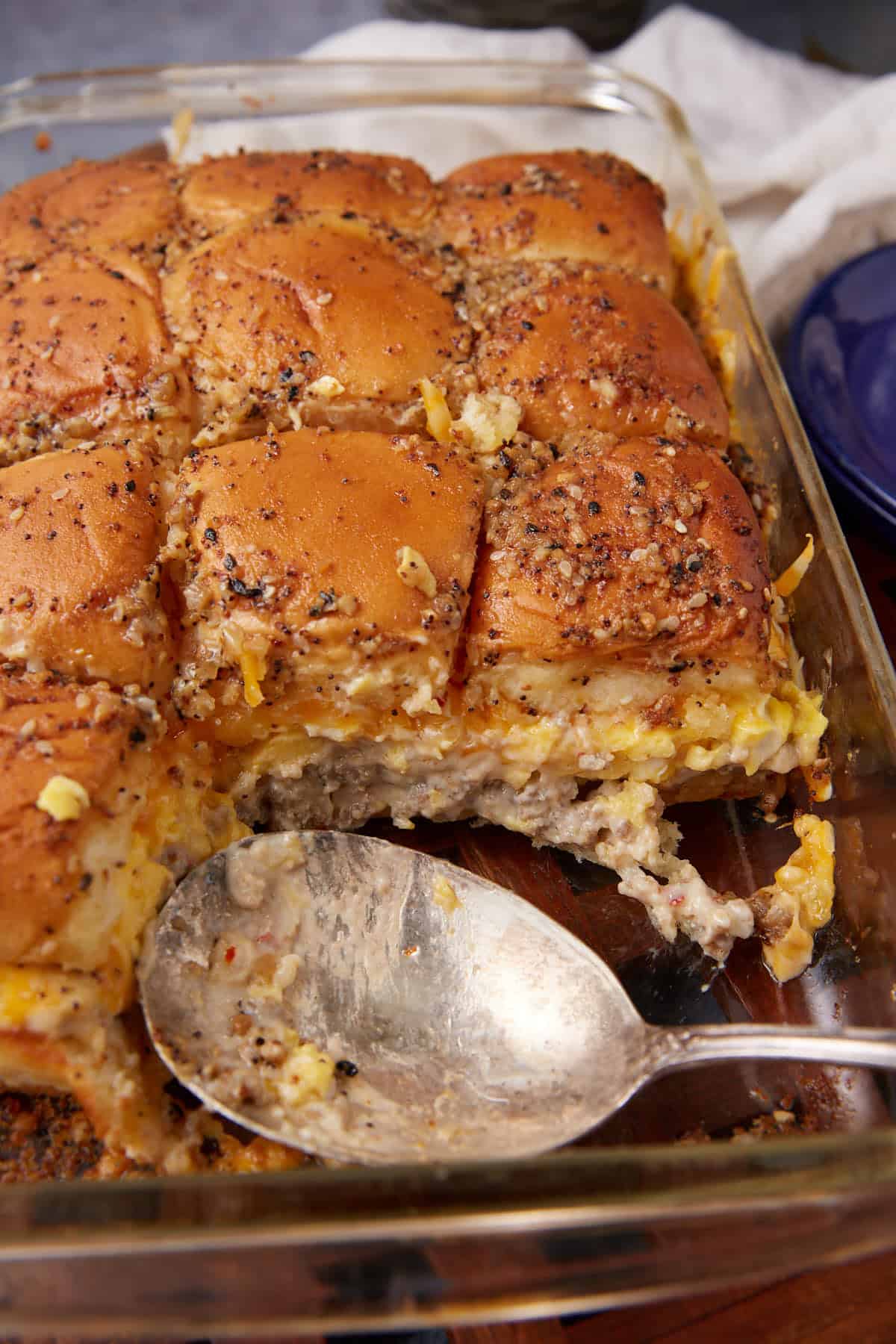 A glass baking dish filled with cheesy breakfast sliders made with golden brown buns and a sausage patty, reminiscent of a hearty German apple pancake breakfast, with a serving spoon and some sliders already removed.