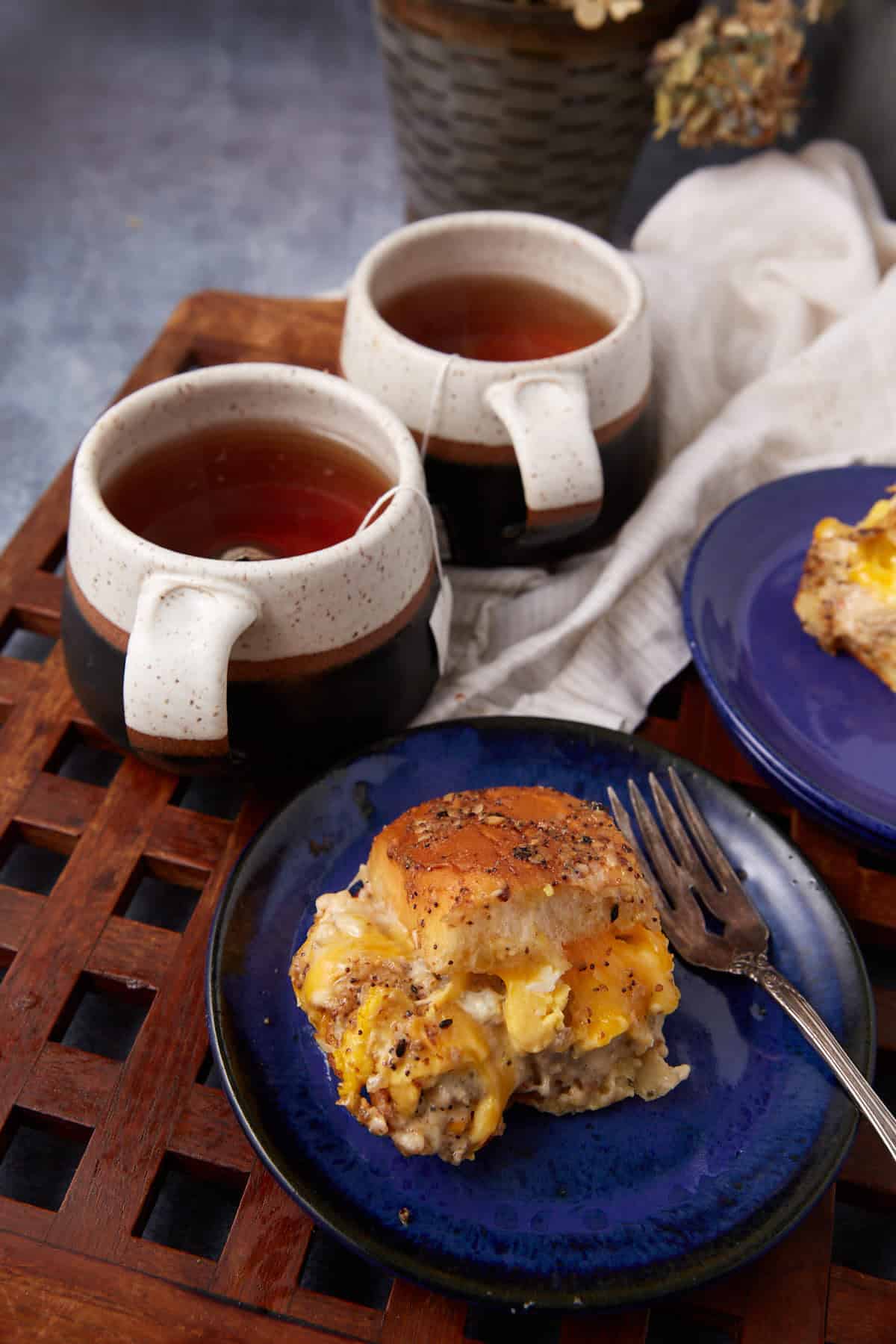 A blue plate holds a biscuit breakfast sandwich with eggs, cheese, and sausage, next to a fork. Two mugs of tea and a German apple pancake sit behind the plate on a wooden tray, with a light cloth and potted plant in the background.
