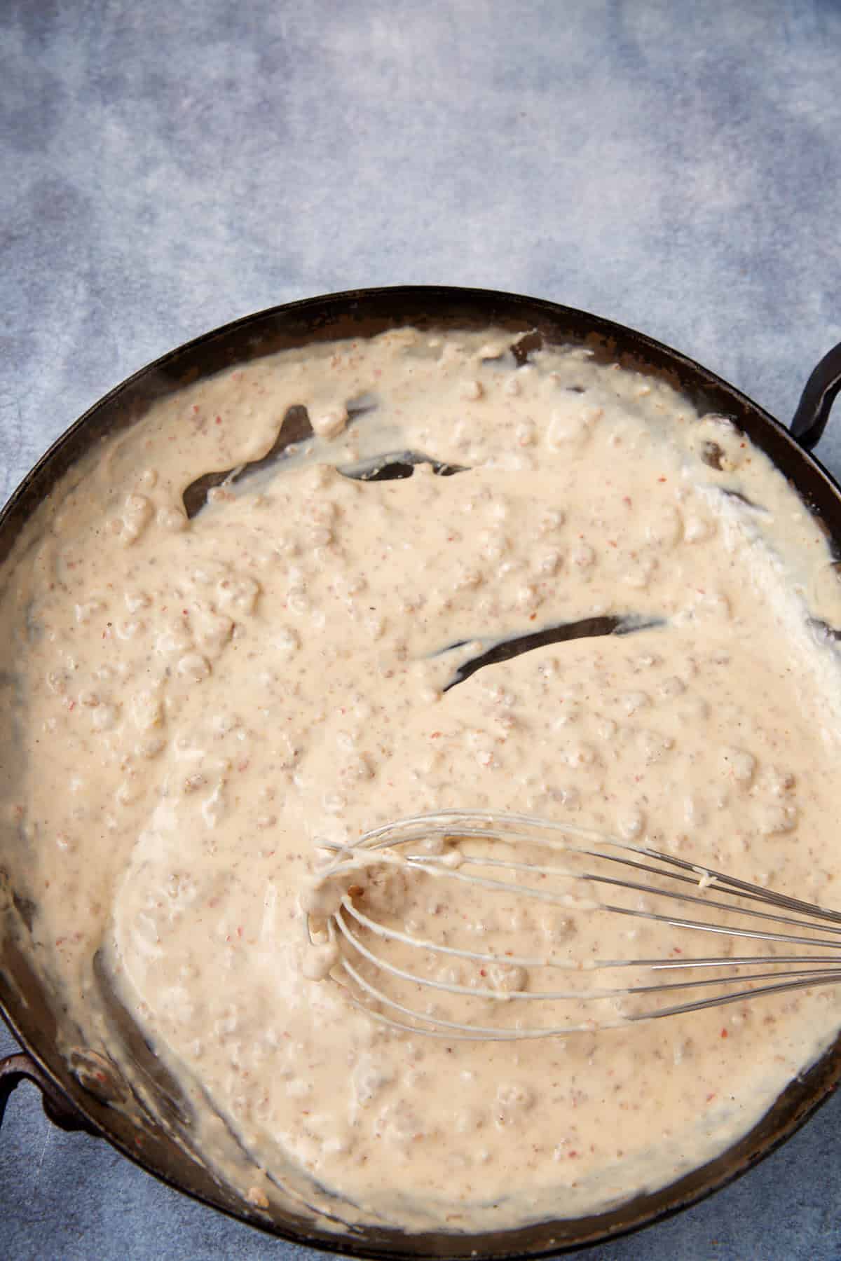 A black skillet filled with creamy sausage gravy being stirred with a metal whisk, set on a light blue textured surface, next to a warm German apple pancake.