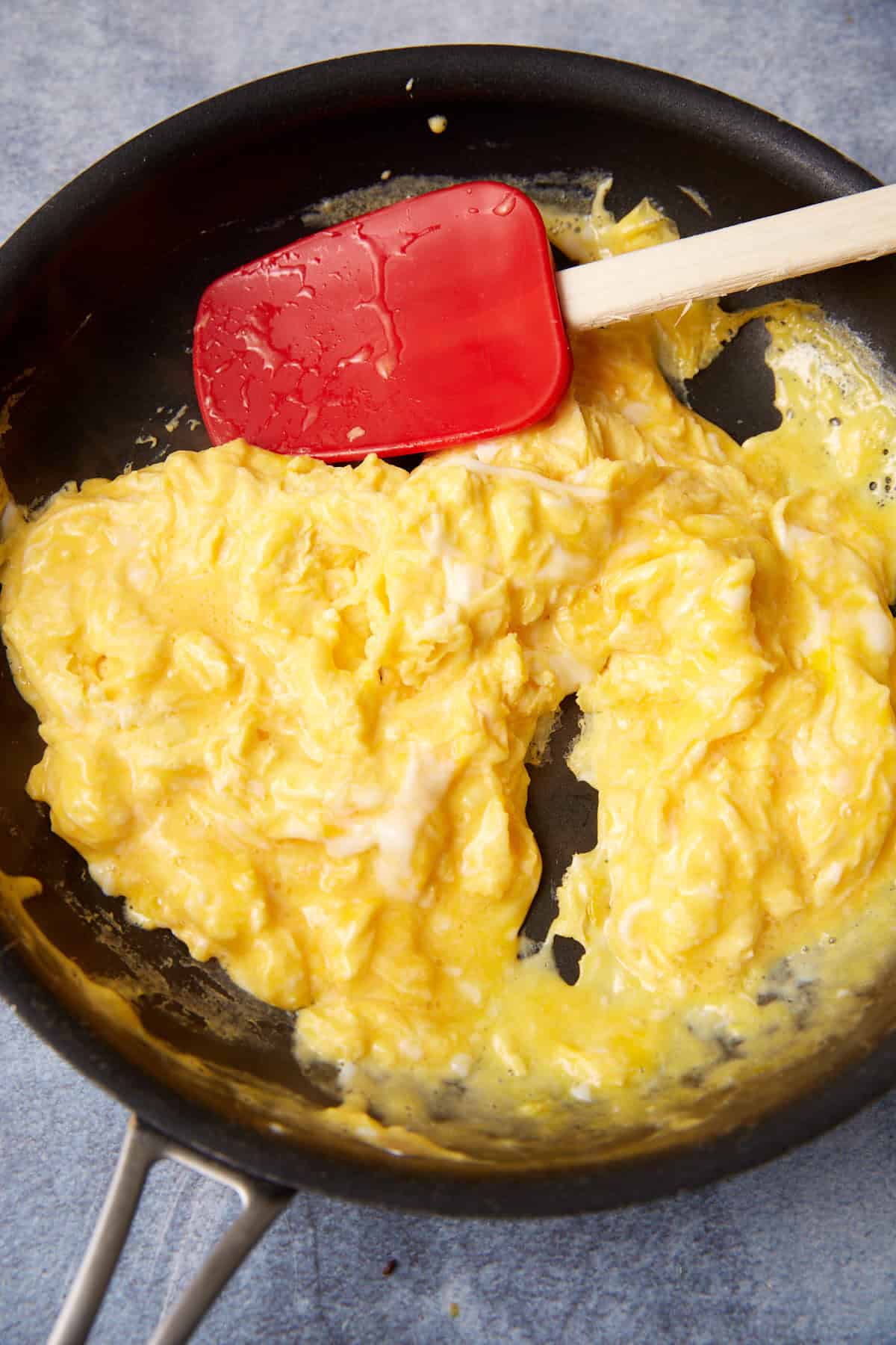 Scrambled eggs being cooked in a black nonstick pan with a red spatula resting on the eggs. The eggs appear soft and creamy, reminiscent of the custardy texture found in a German apple pancake.