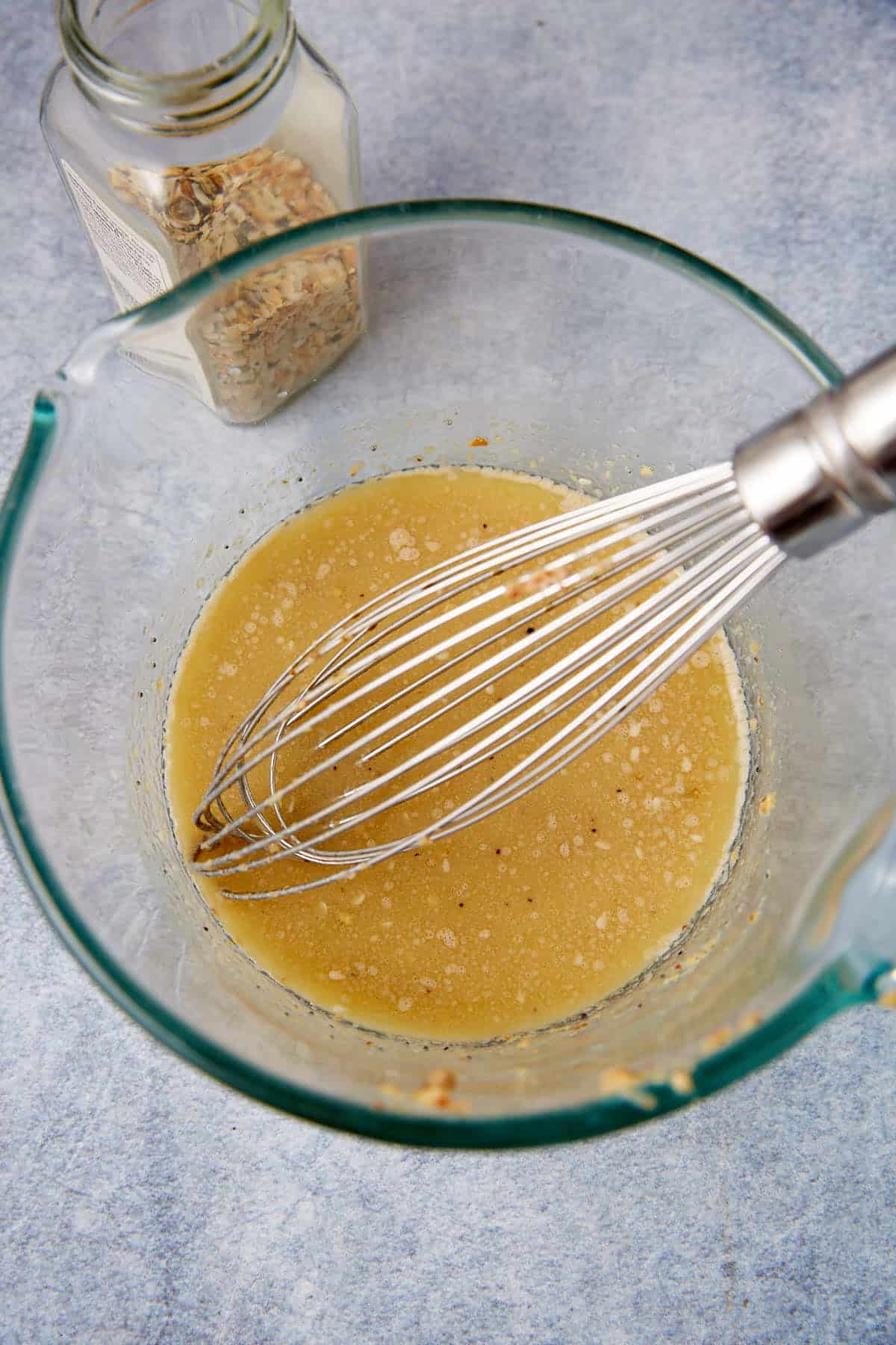 A glass mixing bowl with a metal whisk blending a yellowish batter—perfect for making German apple pancake—sits next to an open jar of oats on a light gray surface.