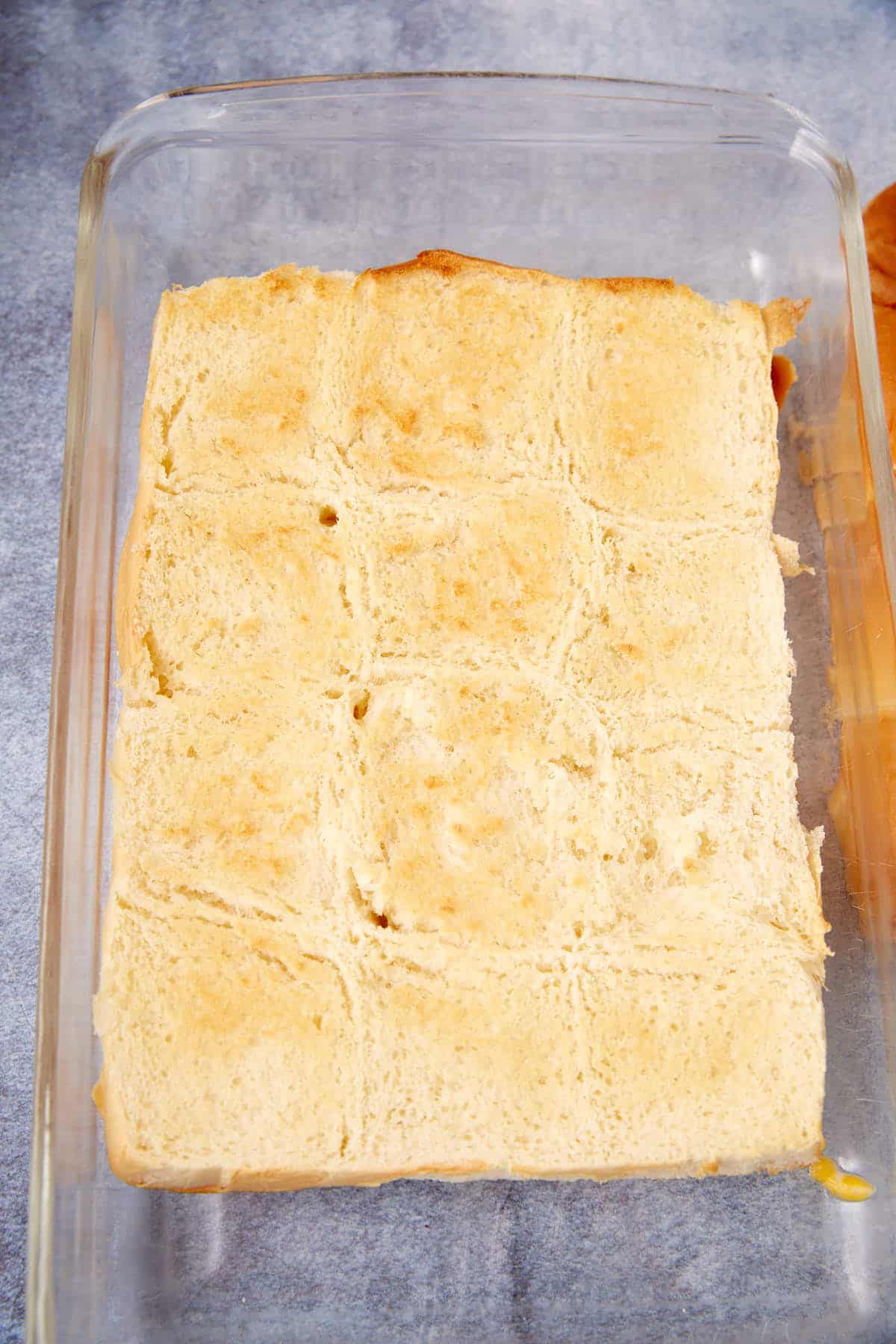 A glass baking dish containing baked, golden-brown slider rolls aligned in a grid pattern with some uneven edges, reminiscent of the rustic look of a German apple pancake, placed on a gray surface.