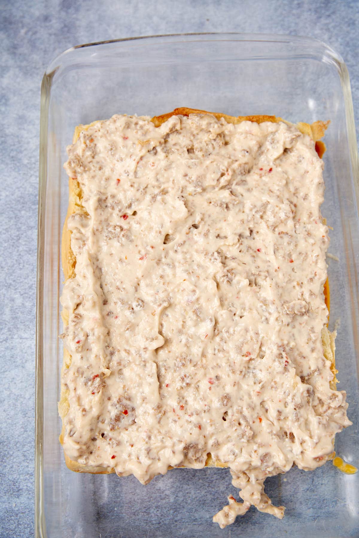 A glass baking dish with a layer of creamy sausage gravy spread over baked biscuit dough, ready for the next step in preparation—reminiscent of the comforting layers found in a German apple pancake. The background is a light gray surface.