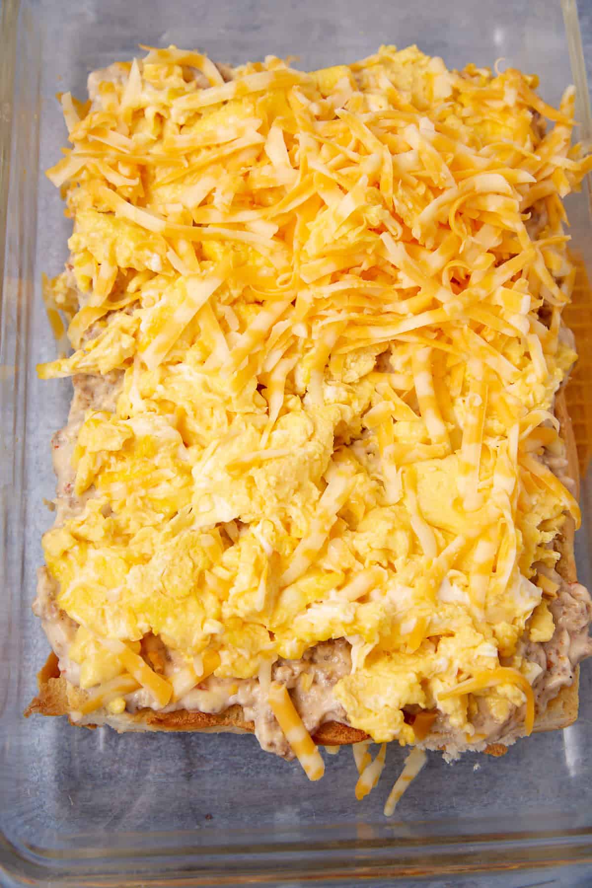 A close-up of a casserole dish filled with layers of scrambled eggs, shredded yellow and white cheese, and a creamy mixture, reminiscent of the comfort found in a German apple pancake, ready to be baked.