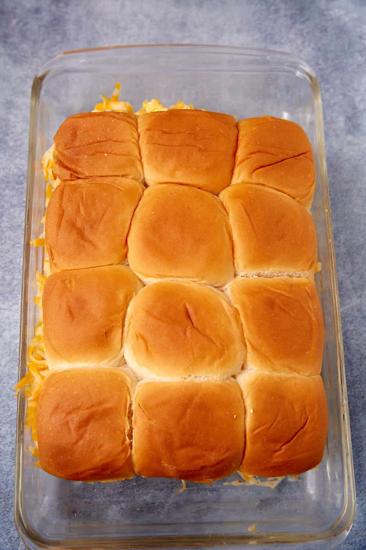 A glass baking dish filled with twelve golden-brown dinner rolls, neatly arranged in a 3x4 grid, with some shredded cheese visible peeking out from the sides—reminiscent of the rustic comfort of a German apple pancake.