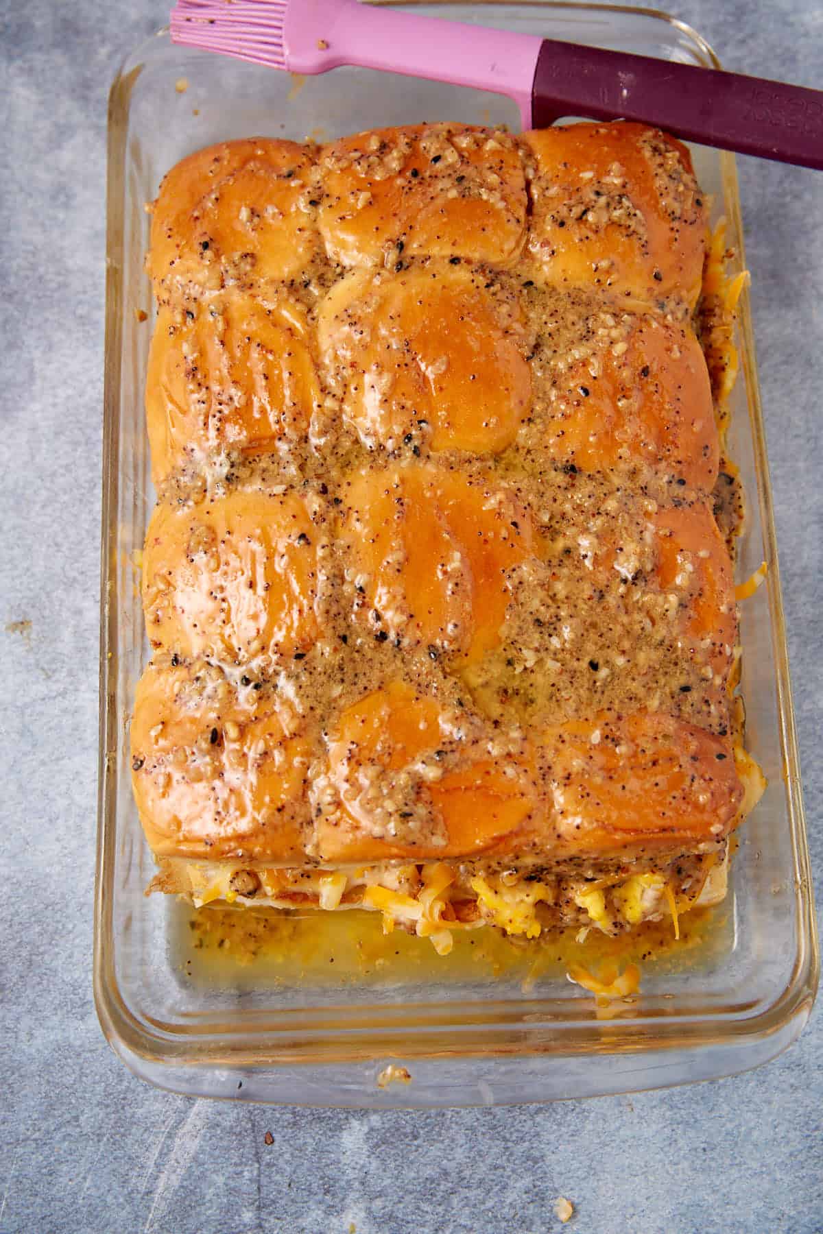 A glass baking dish filled with golden, seasoned slider buns, topped with a glaze and visible melted cheese—reminiscent of the tempting finish on a classic German apple pancake—with a pink and purple pastry brush resting on the edge.