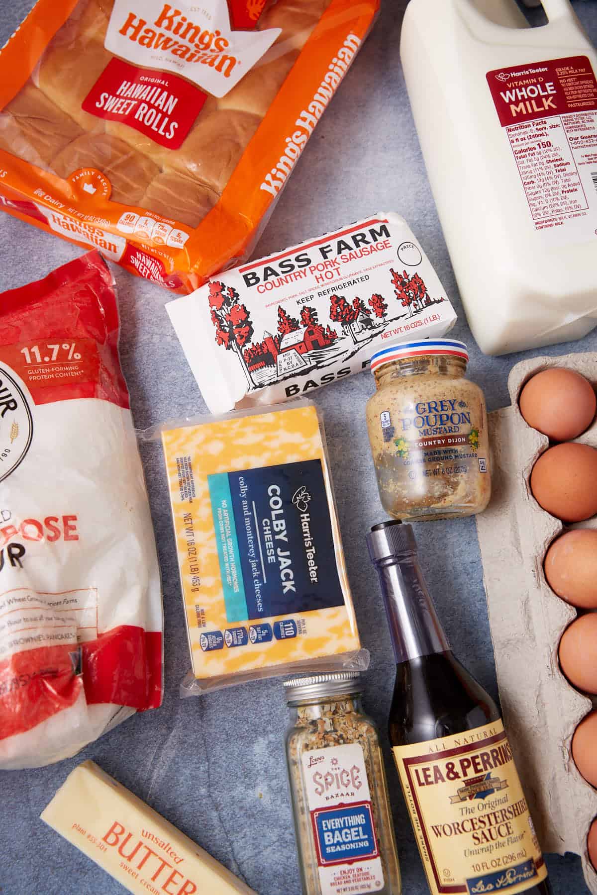 An assortment of groceries on a counter, including Hawaiian sweet rolls, whole milk, eggs, flour for a German apple pancake, butter, colby jack cheese, country pork sausage, Worcestershire sauce, two types of seasoning, and a jar of coarse mustard.