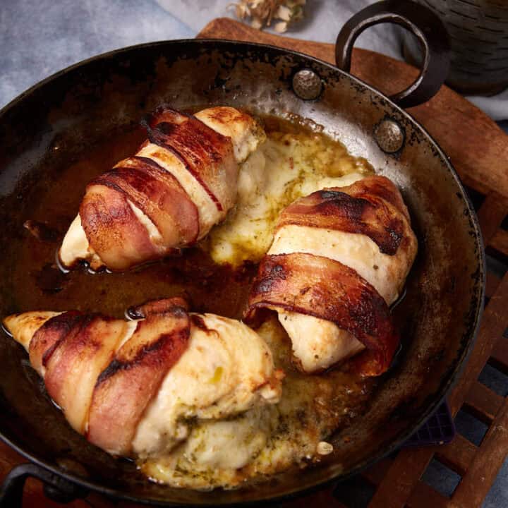 Three mozzarella stuffed chicken breasts wrapped in crispy bacon are cooked in a rustic metal skillet, with melted cheese and juices pooling around the chicken. The dish appears golden brown and flavorful.