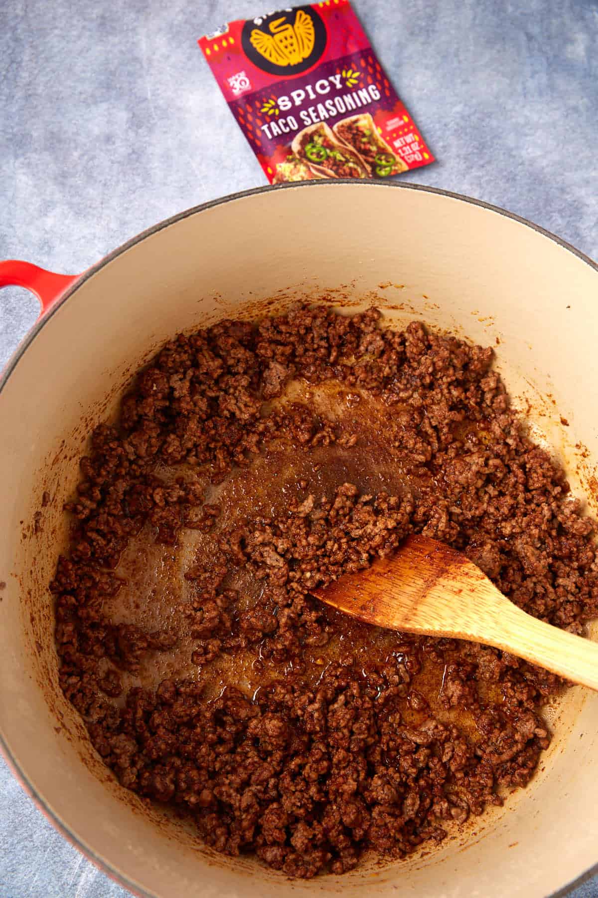Cooked ground beef mixed with spicy taco seasoning simmers in a large pot, stirred with a wooden spoon—an easy taco spaghetti casserole perfect for busy nights. A packet labeled “Spicy Taco Seasoning” sits in the background on a blue surface.