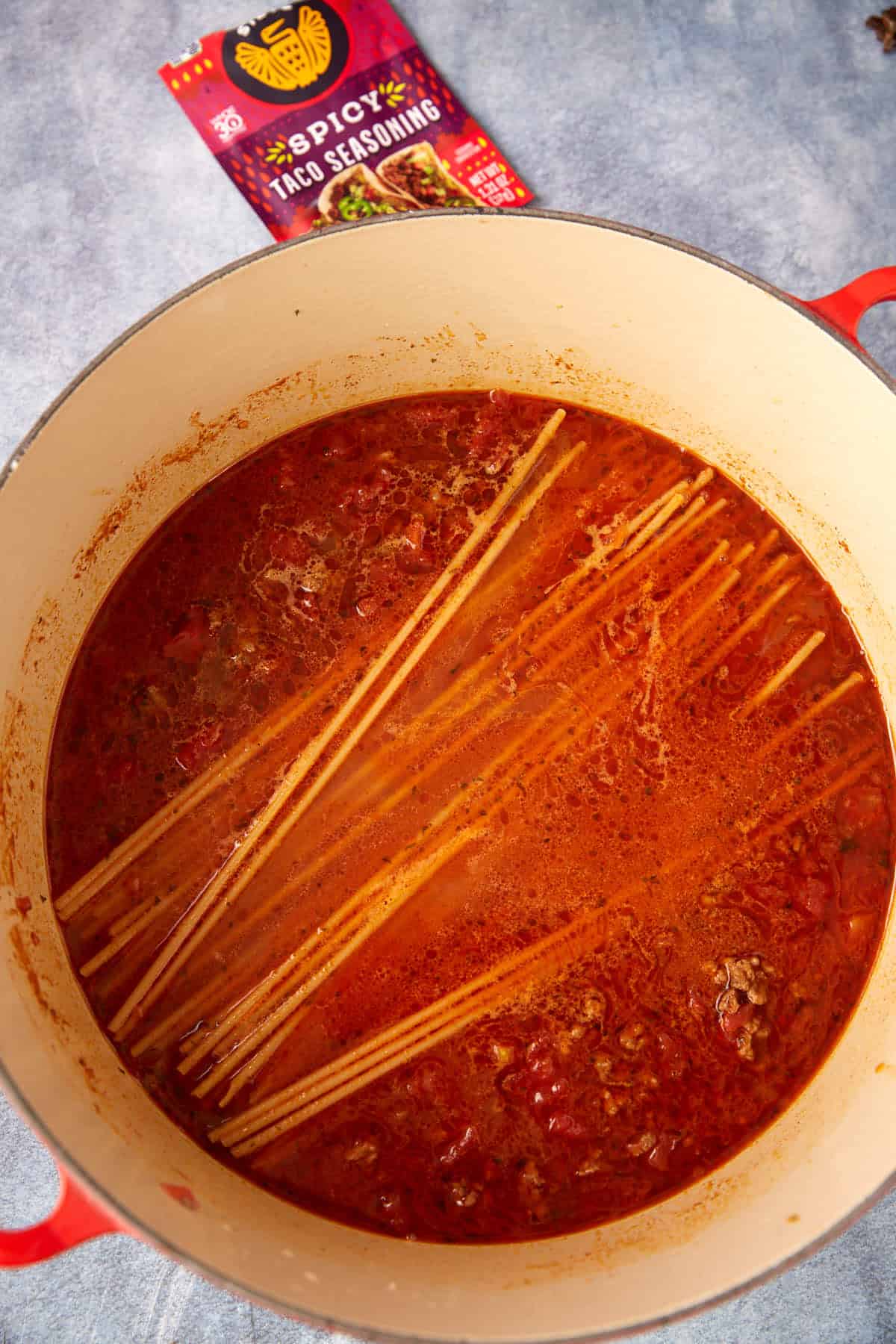 A large pot filled with a red, tomato-based sauce, partially submerged uncooked spaghetti, and bits of ground meat hints at a tasty one pot taco spaghetti. A packet of spicy taco seasoning sits in the background on a gray surface.