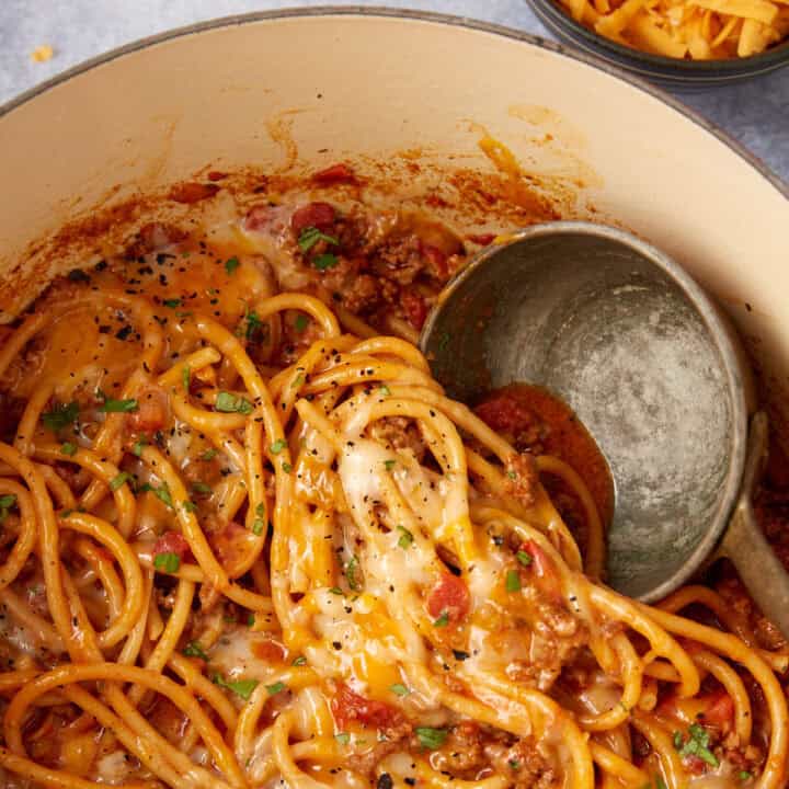 A close-up of a pot filled with cheesy taco spaghetti one pot, ground meat, and tomato sauce, topped with melted cheese and chopped herbs. A metal ladle rests in the pot, ready to serve this delicious taco spaghetti recipe.