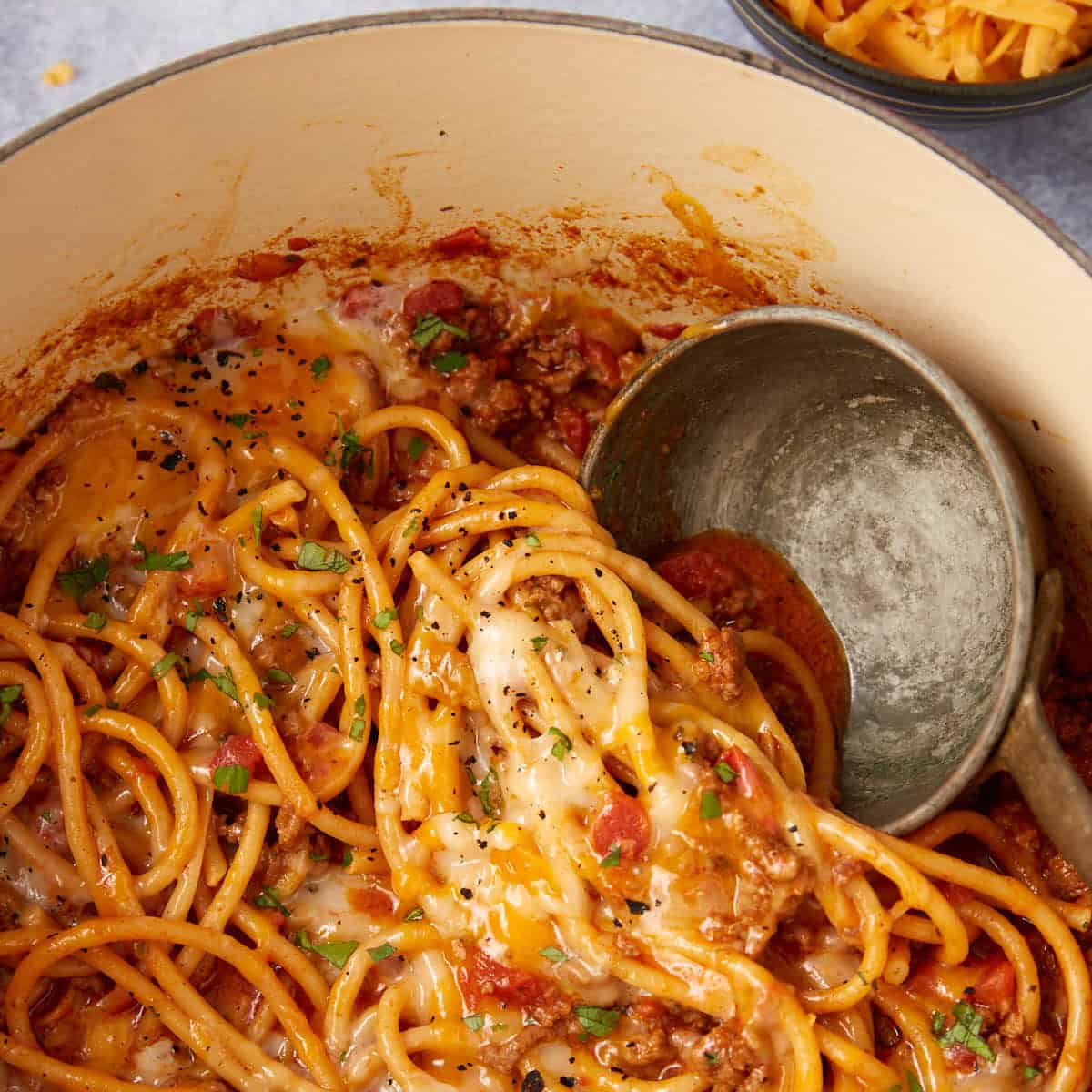 A close-up of a pot filled with cheesy taco spaghetti one pot, ground meat, and tomato sauce, topped with melted cheese and chopped herbs. A metal ladle rests in the pot, ready to serve this delicious taco spaghetti recipe.