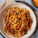 A plate of taco spaghetti casserole with a rich meat sauce, topped with melted cheddar cheese and garnished with chopped herbs, is served on a white dish over a beige napkin.