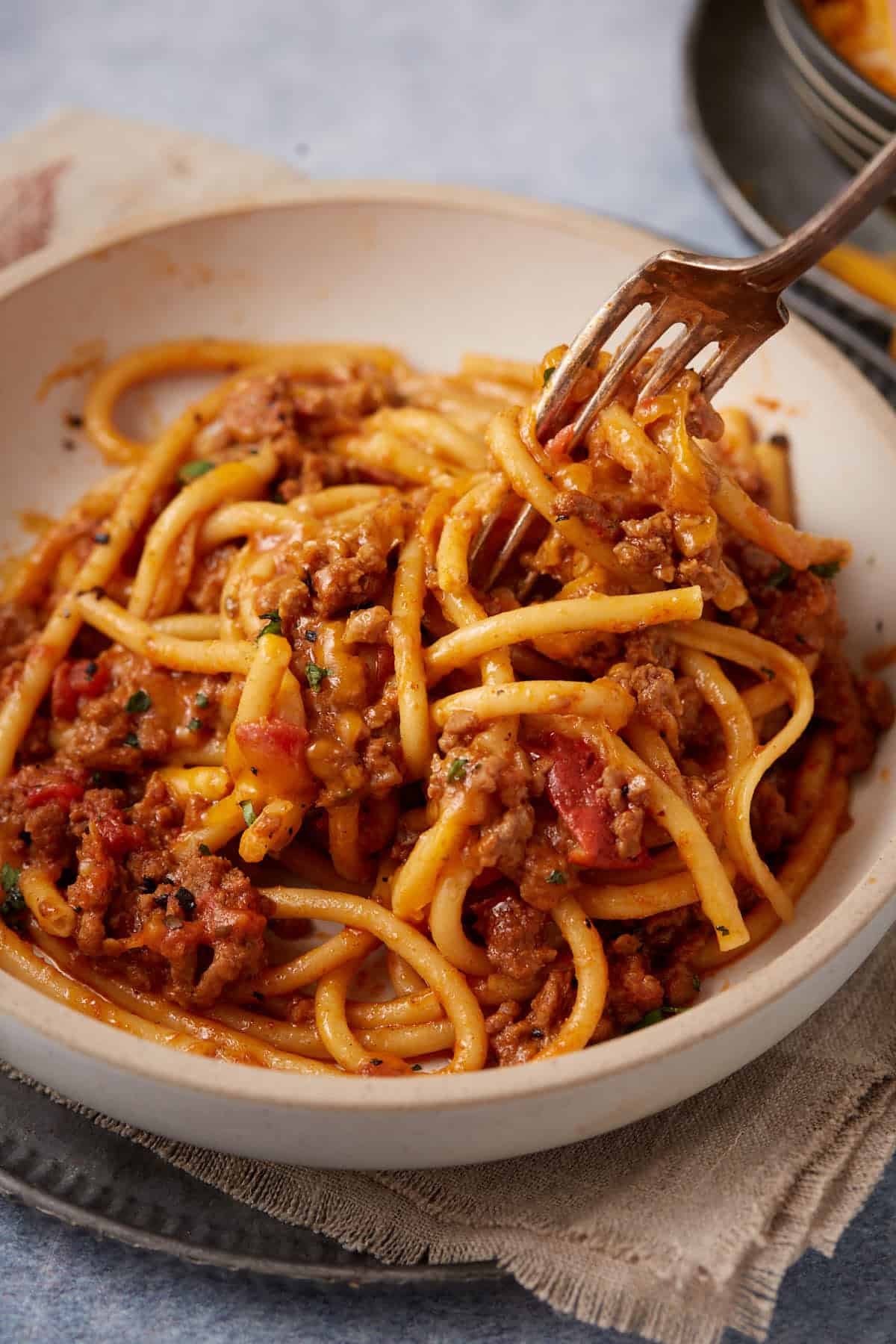A fork twirls thick spaghetti noodles coated in a rich meat sauce with ground beef, tomatoes, and herbs—just like an easy taco spaghetti—served in a shallow white bowl on a beige napkin.