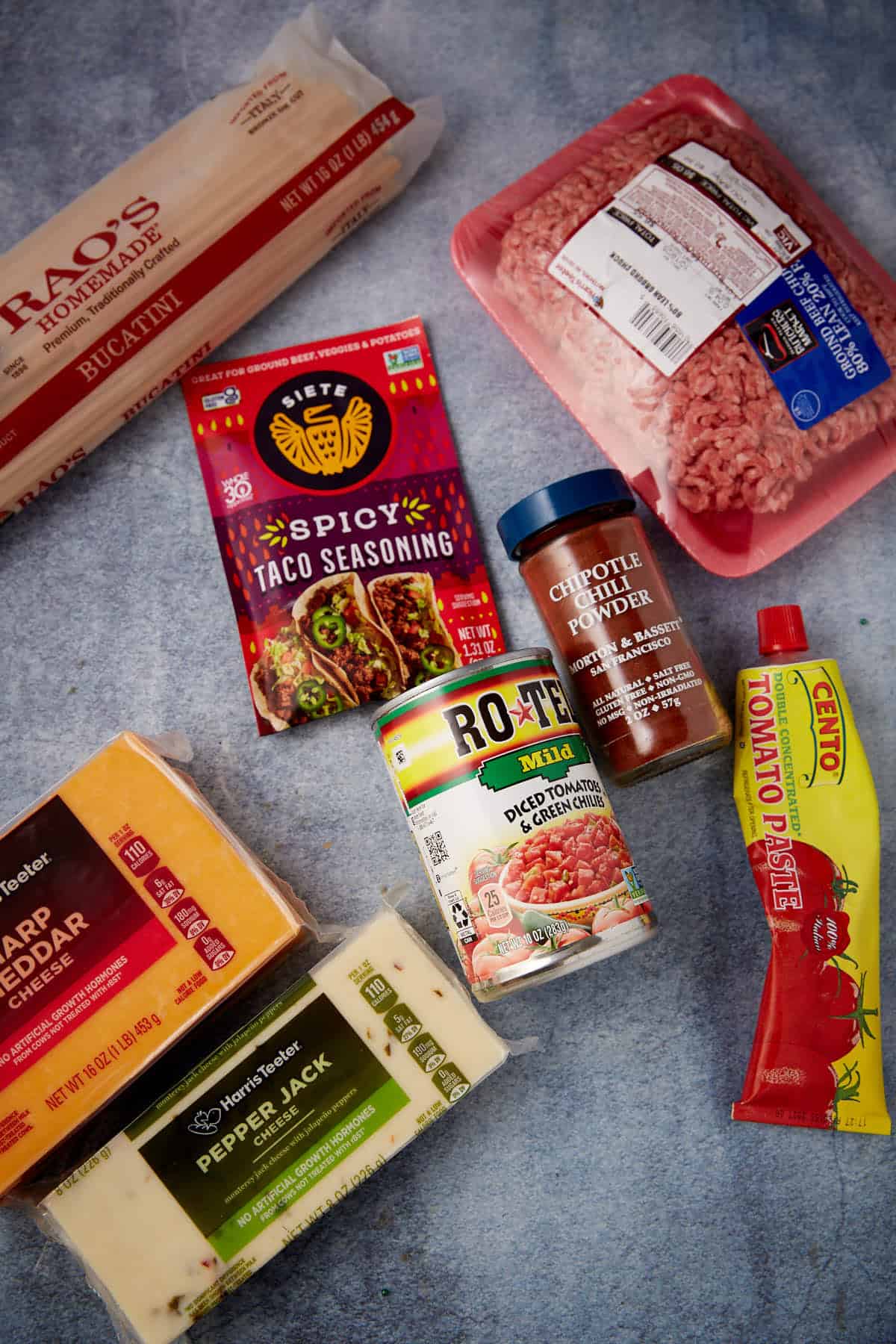 Assorted ingredients on a blue surface, perfect for one pot taco spaghetti—featuring bucatini pasta, ground beef, sharp cheddar and pepper jack cheese, spicy taco seasoning, chipotle chili powder, Ro-Tel tomatoes, and tomato paste.