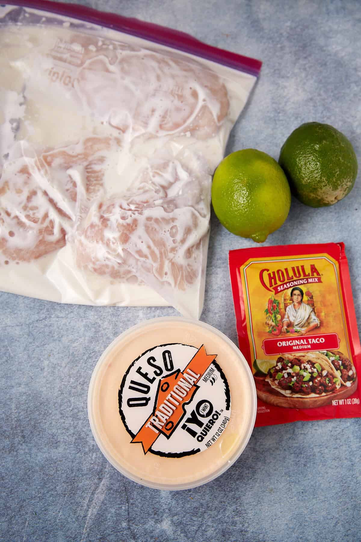 A bag of marinating taco chicken, two whole limes, a packet of Cholula taco seasoning mix, and a container of traditional queso sit on a blue surface.