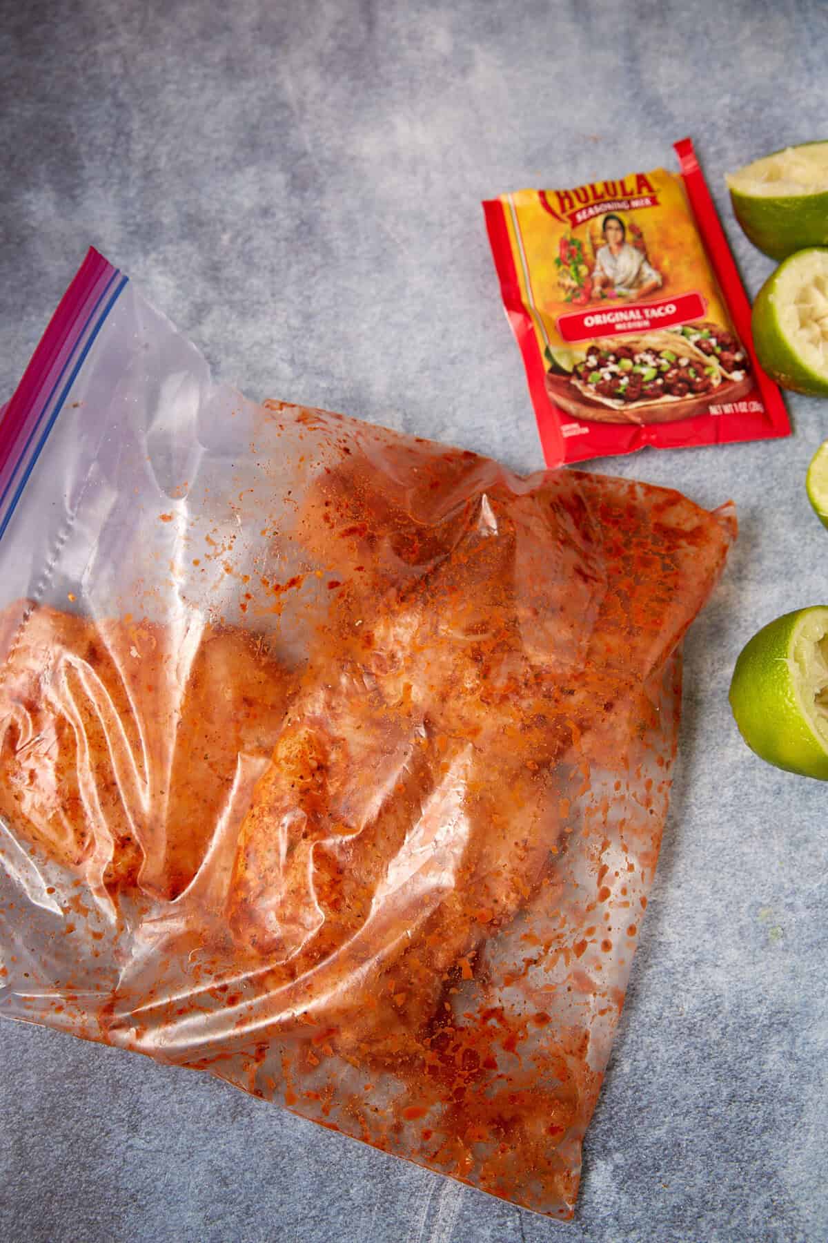 Taco Chicken marinating in a resealable plastic bag with red seasoning, alongside a packet of Cholula Original Taco seasoning and sliced limes on a blue surface.