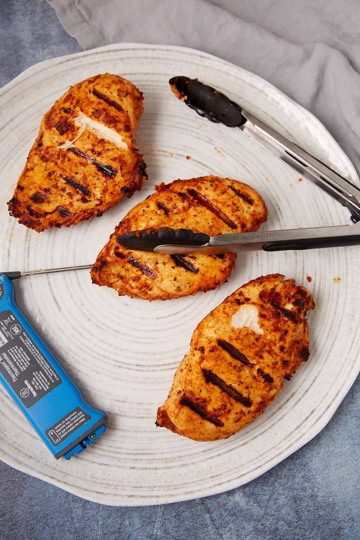 Three Taco Chicken breasts, perfectly grilled with visible char marks, rest on a white plate. Two metal tongs and a blue meat thermometer lie beside them, with a gray cloth nearby.