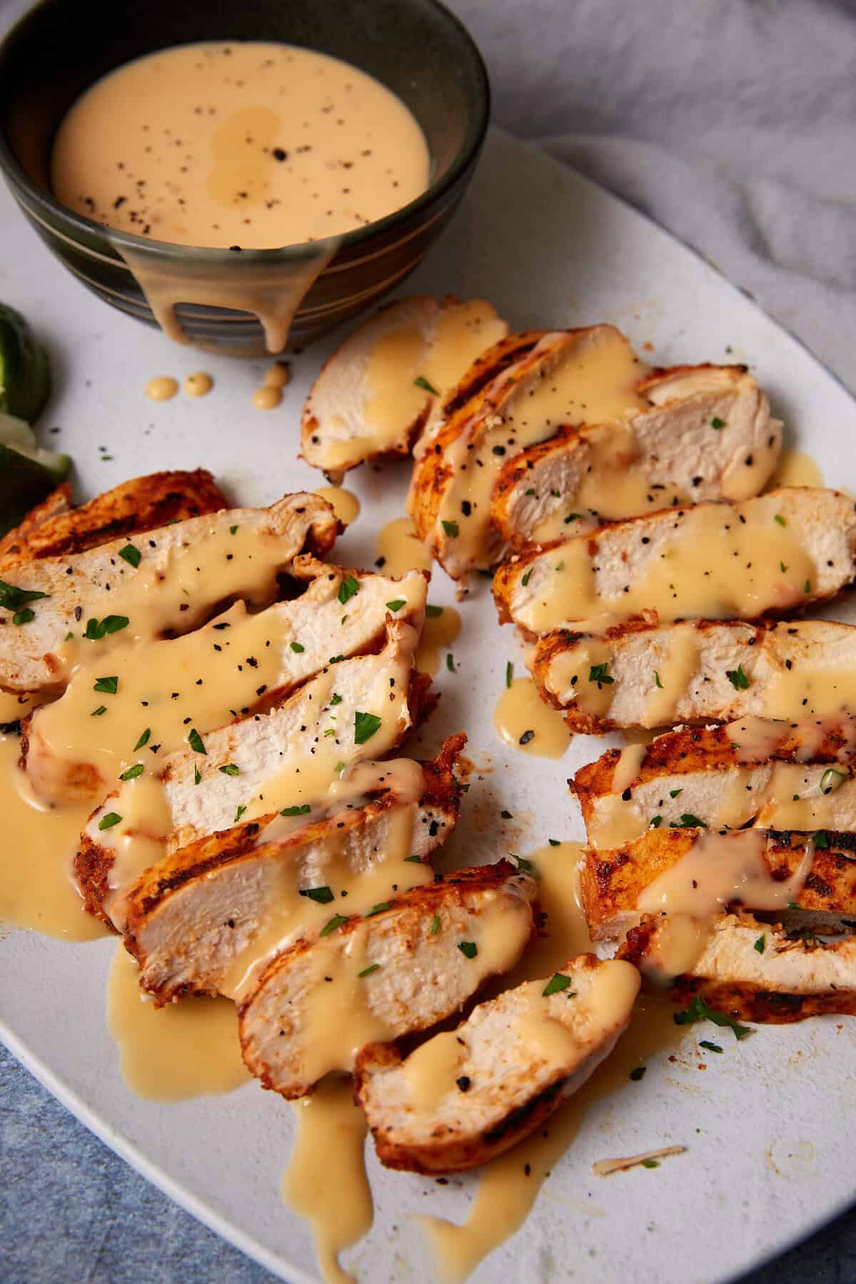 Sliced Taco Chicken breast drizzled with creamy cheese sauce and garnished with chopped herbs, served on a white plate with a bowl of extra sauce in the background.