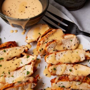 Sliced grilled taco chicken breast topped with creamy yellow sauce and sprinkled with herbs and black pepper, served beside a small bowl of extra sauce, with a black fork nearby.