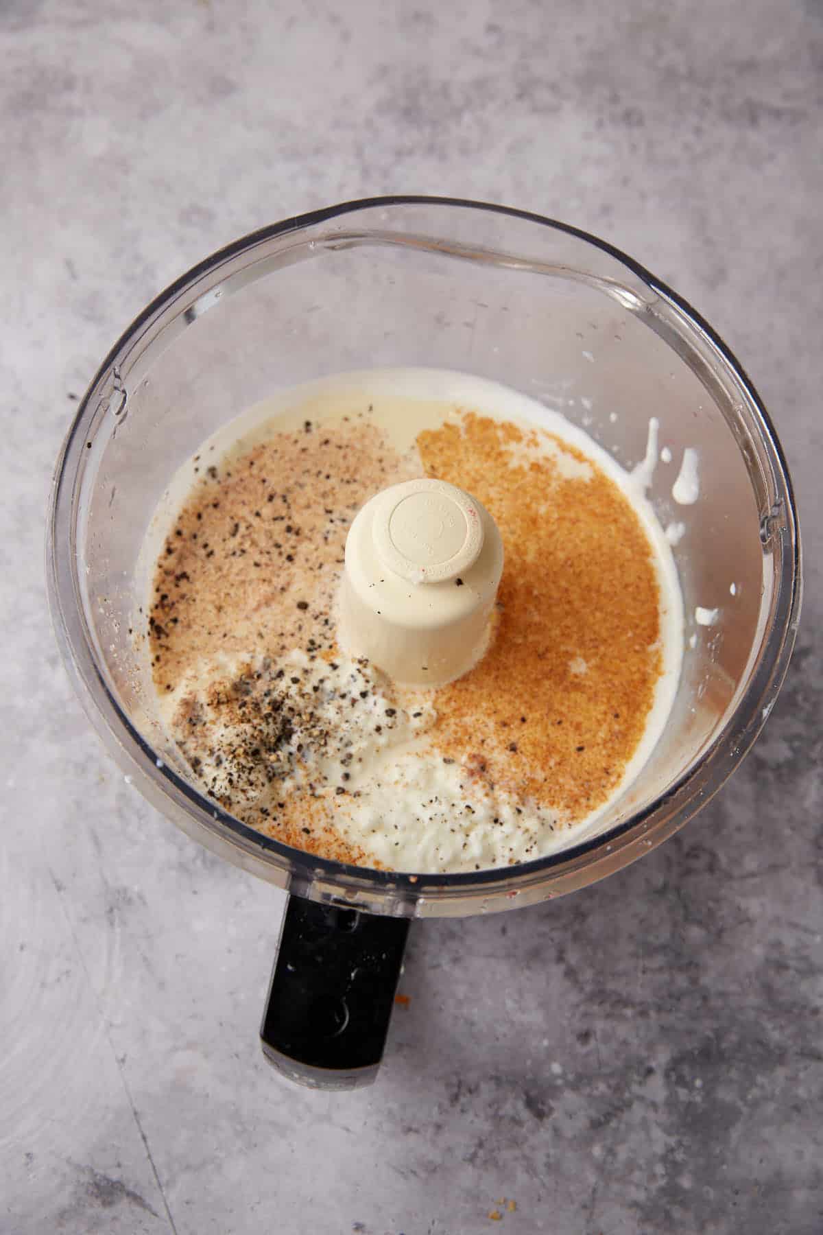 A food processor bowl containing cream, breadcrumbs, and cracked black pepper—ideal ingredients for topping your favorite One Pot Macaroni and Cheese—ready to be blended on a gray countertop.