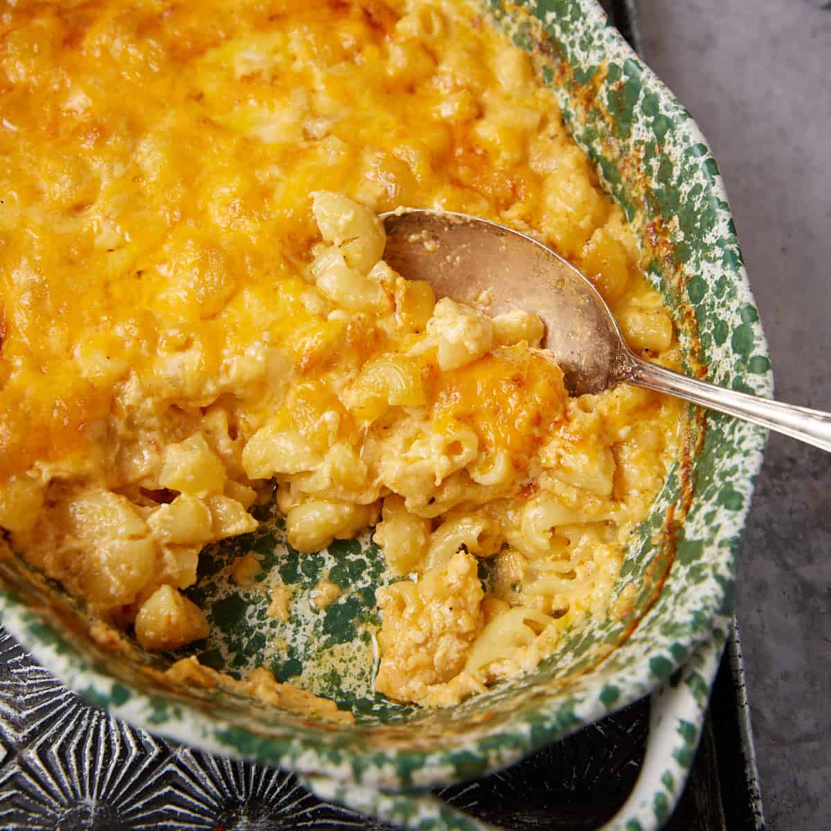 A baked One Pot Macaroni and Cheese casserole in a green and white ceramic dish, partially served with a metal spoon, reveals creamy pasta beneath a golden, melted cheese topping.