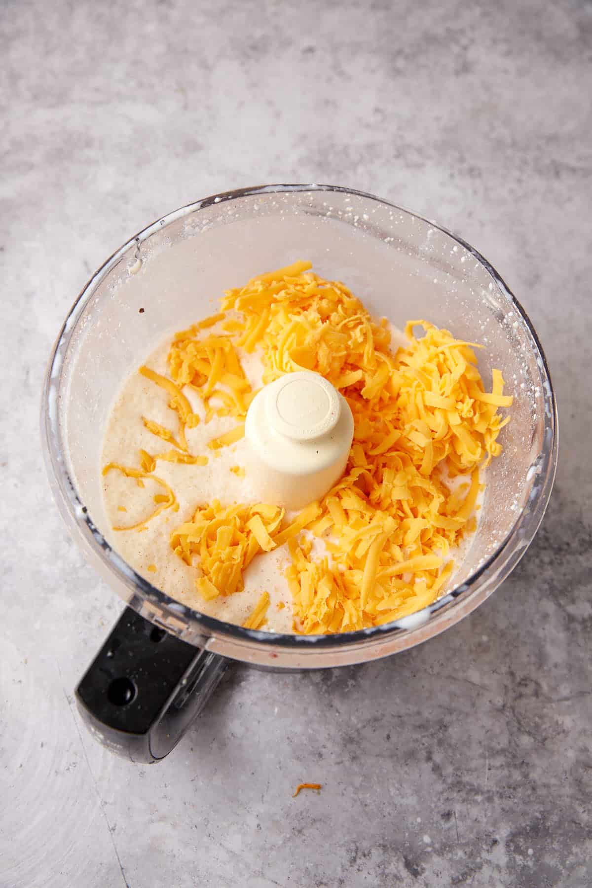 A food processor bowl containing grated cheddar cheese and a creamy liquid mixture for One Pot Macaroni and Cheese, placed on a gray countertop.