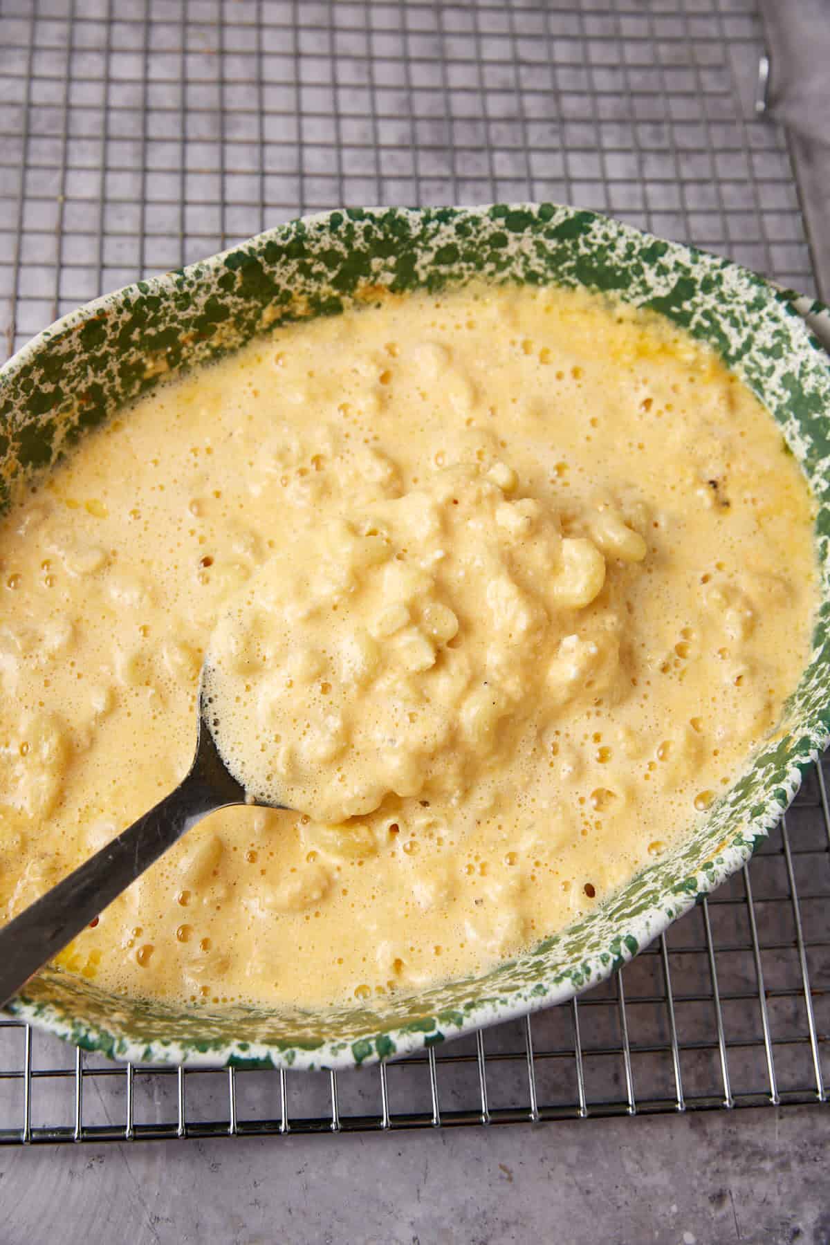 A metal spoon rests in a green, speckled bowl filled with thick, creamy One Pot Macaroni and Cheese batter, sitting on a metal cooling rack over a gray surface.