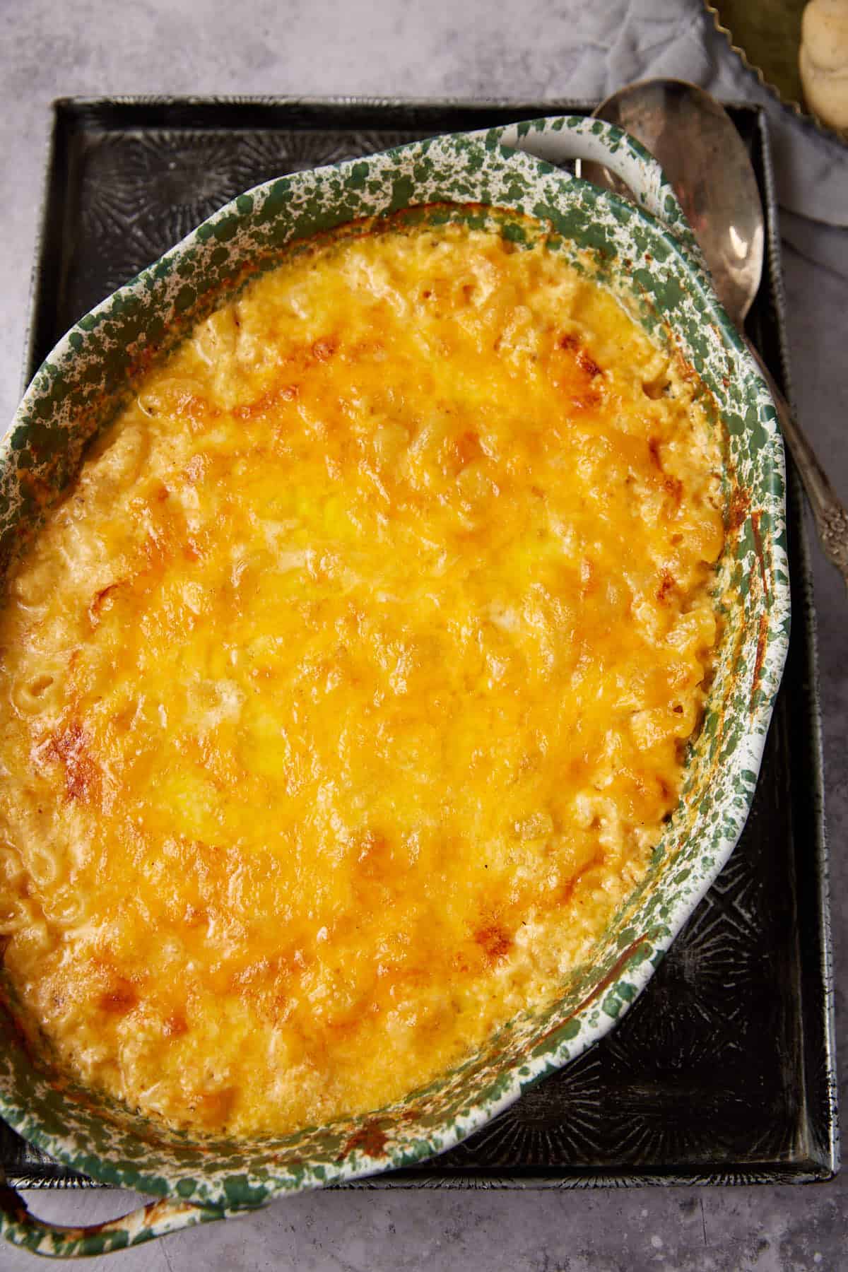 A baked One Pot Macaroni and Cheese casserole in a green-speckled oval dish, topped with melted, golden-brown cheese, sits on a dark tray with a serving spoon beside it.