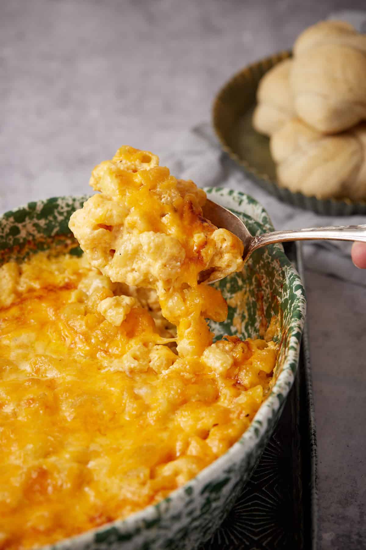 A serving spoon lifts a portion of creamy One Pot Macaroni and Cheese topped with melted cheddar from a green patterned dish, with a plate of dinner rolls in the background.