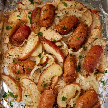 Enjoy a sausage sheet pan dinner with sliced sausages, apple wedges, and onions baked on a foil-lined tray with rice, seasoned with black pepper, and garnished with chopped parsley.