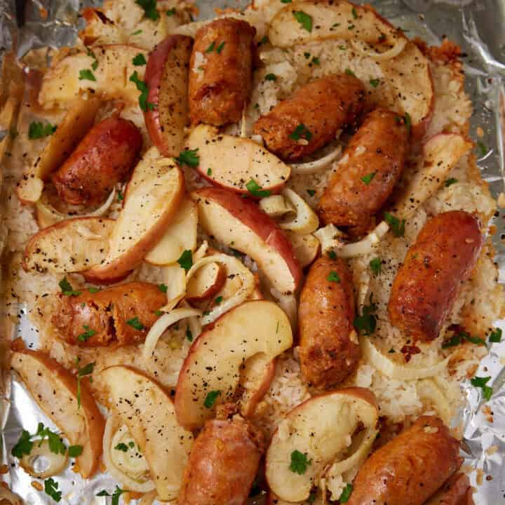 Enjoy a sausage sheet pan dinner with sliced sausages, apple wedges, and onions baked on a foil-lined tray with rice, seasoned with black pepper, and garnished with chopped parsley.